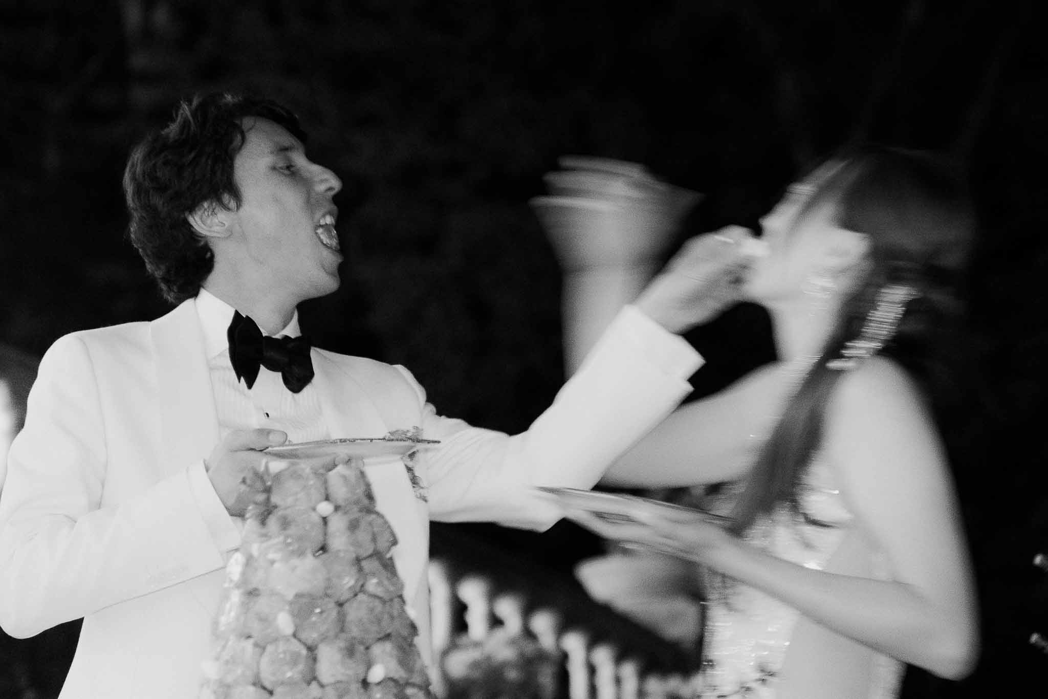 Groom feeds bride cake beside croquembouche tower in candlelit motion-blurred reception moment in B&W