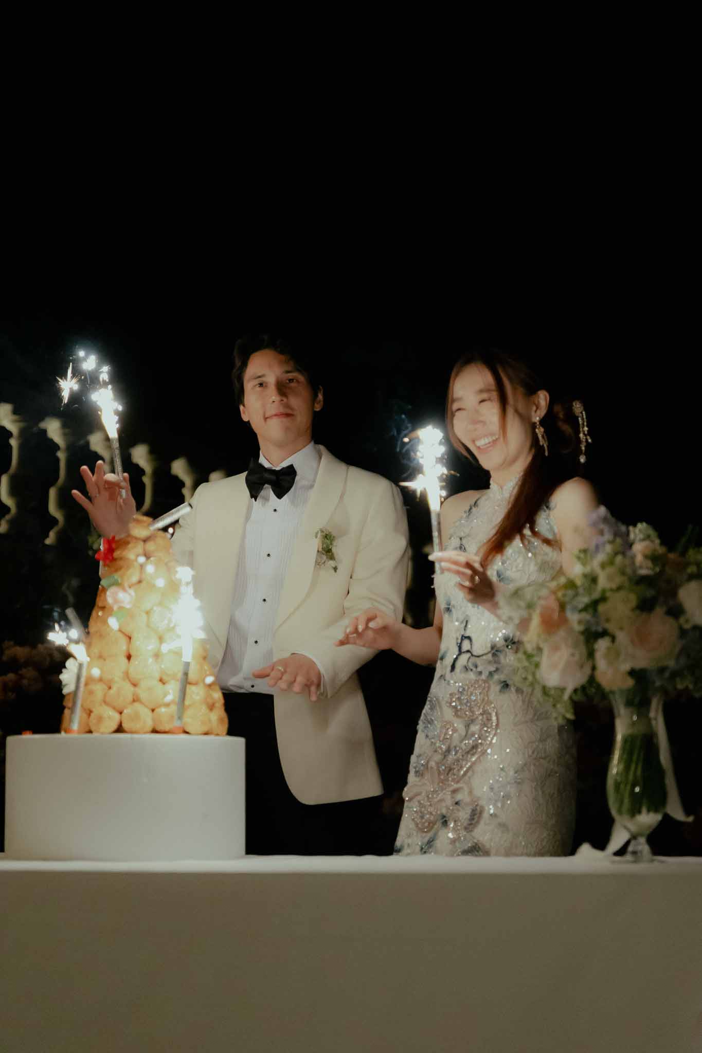 Couple beside sparkler-topped croquembouche with blush rose arrangement at dark outdoor evening reception