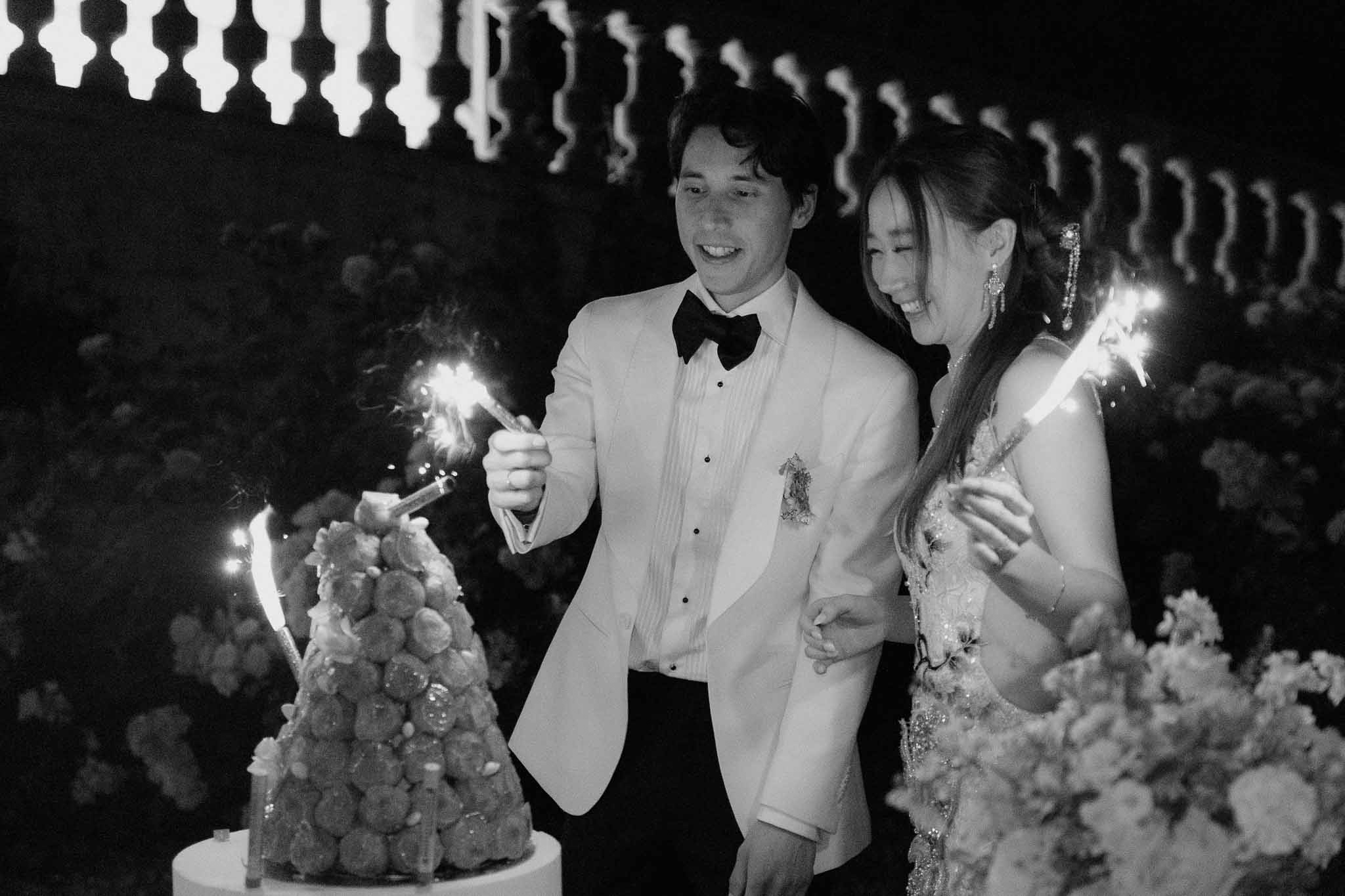Bride and groom cutting a croquembouche with lit sparklers during outdoor evening reception in black and white