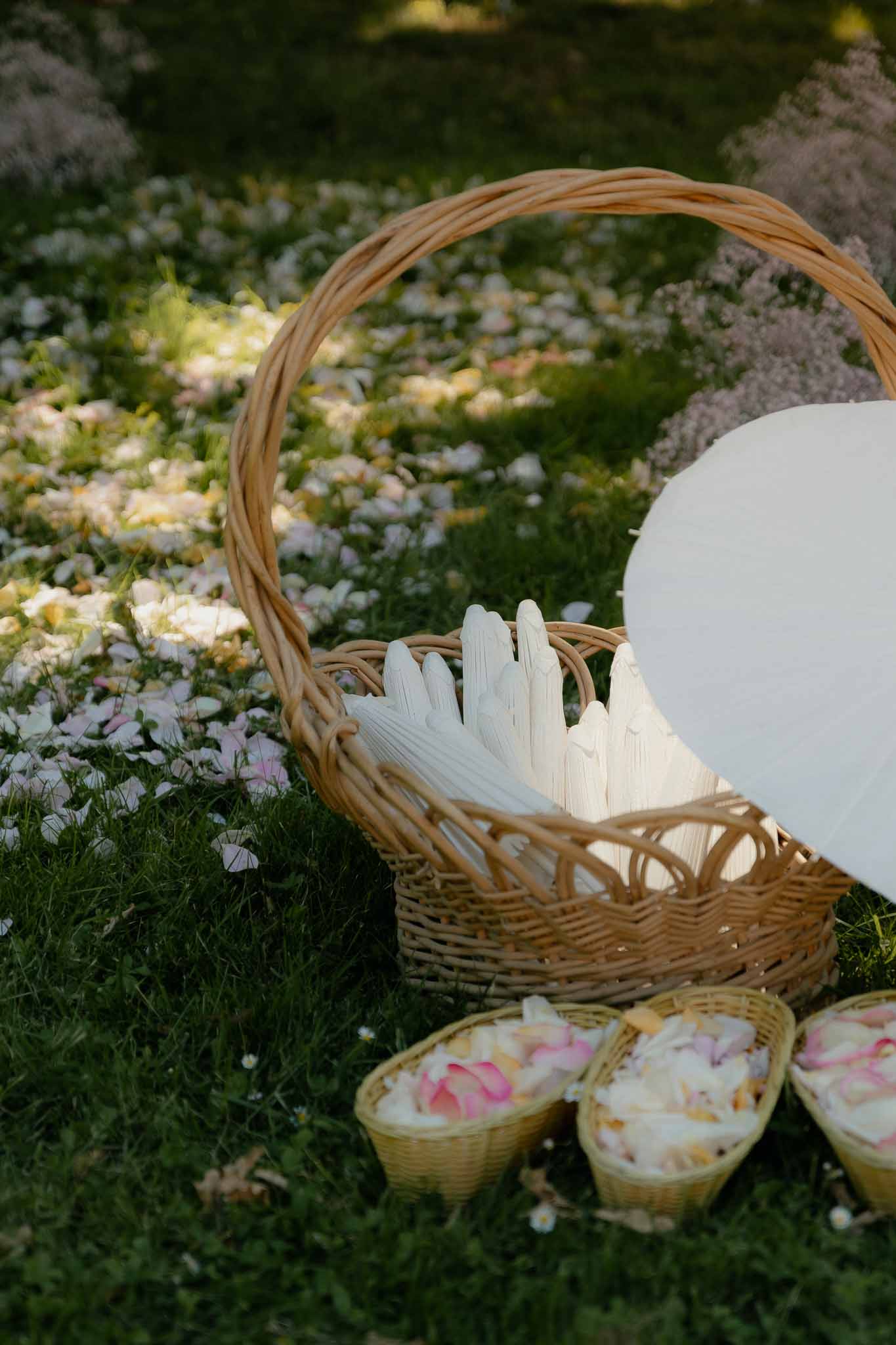 Wicker basket of white paper hand fans and cone holders with pink, white, and yellow flower petals on grass