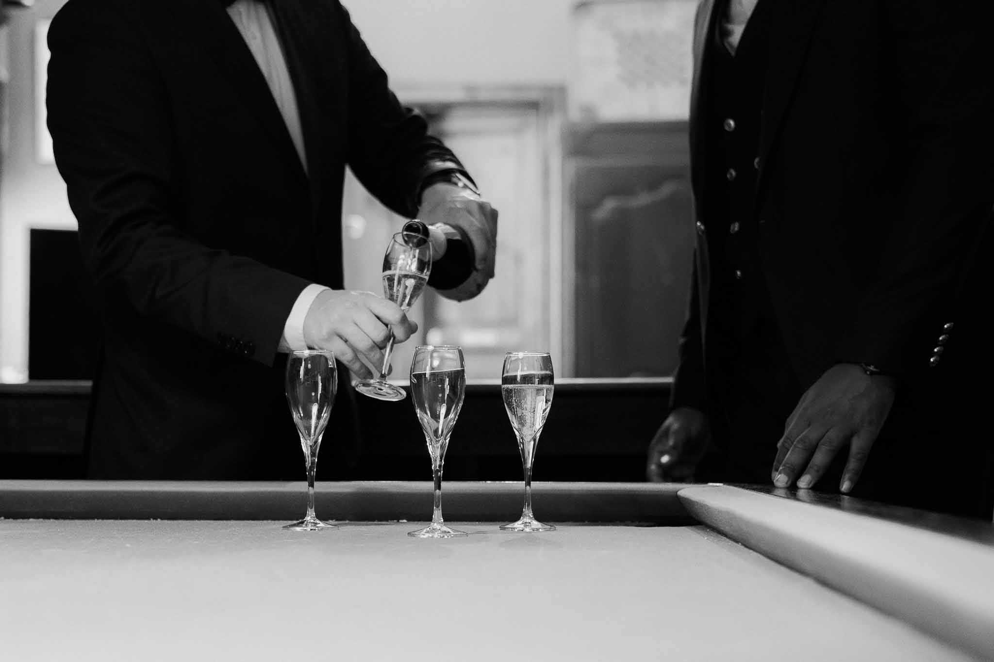 Black and white close-up of champagne being poured into flutes on a bar counter during wedding reception