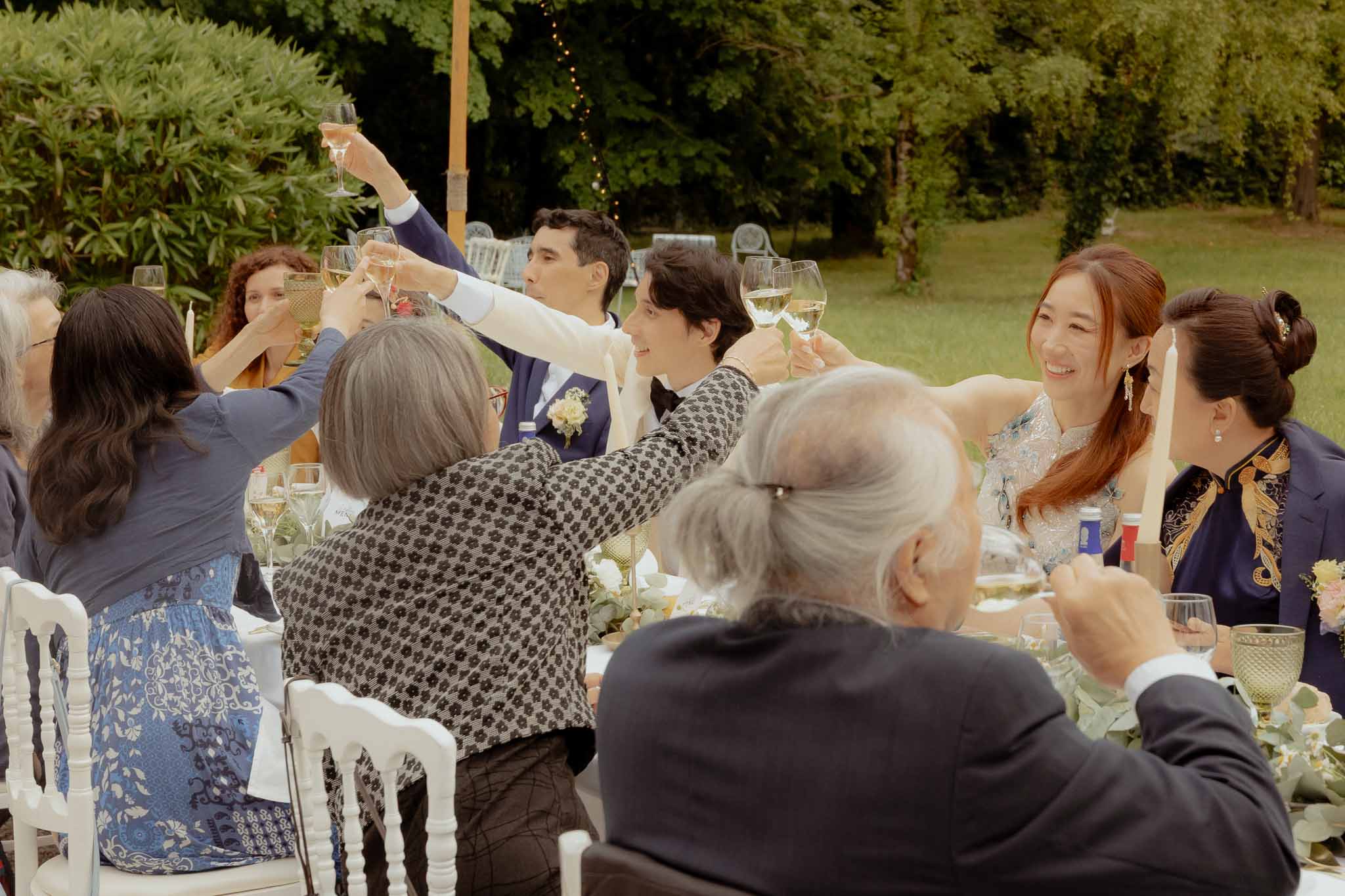 Couple and guests raising glasses for champagne toast at outdoor reception table with fairy lights