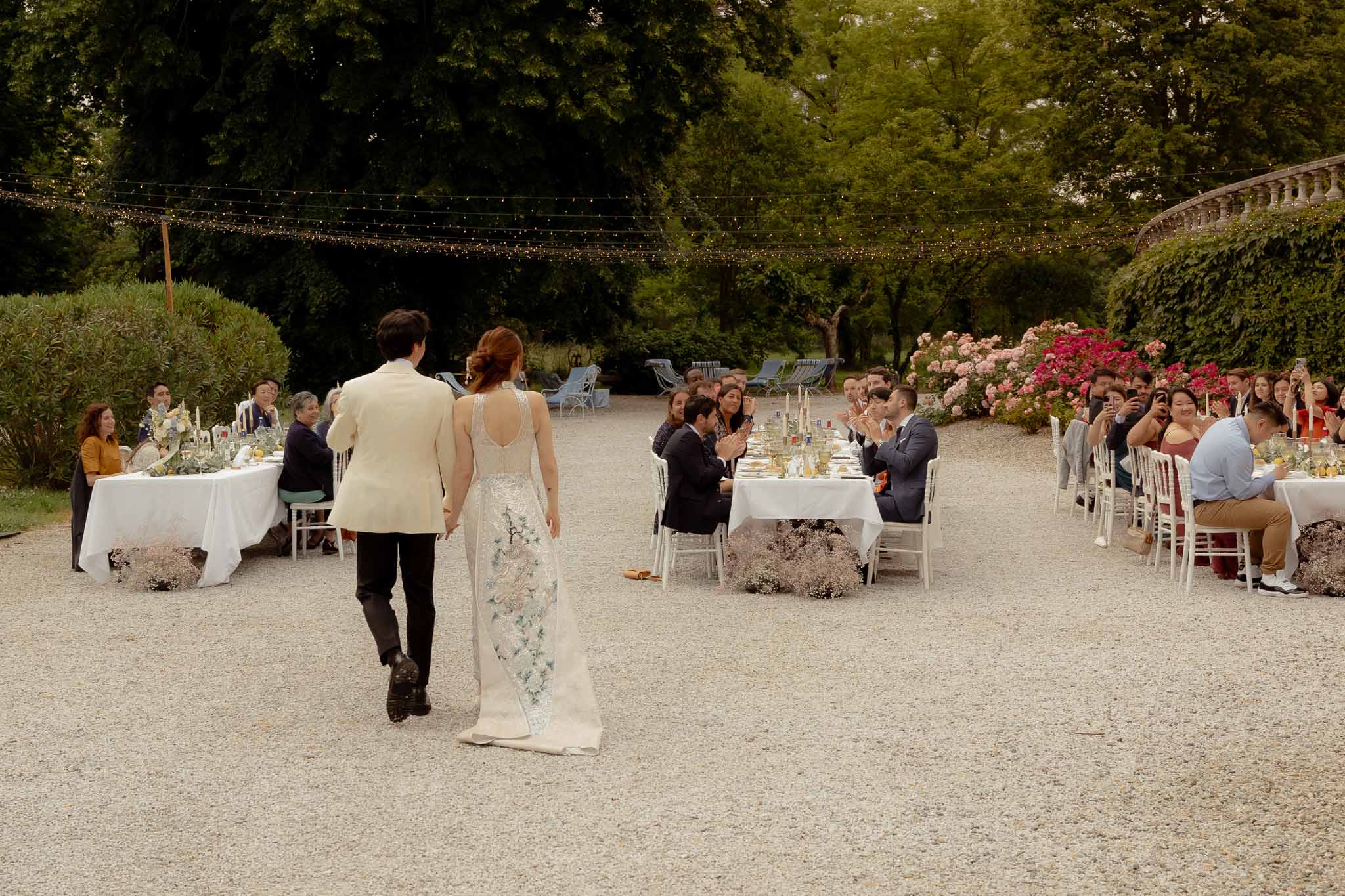Couple entering garden reception between banquet tables as guests applaud under fairy lights
