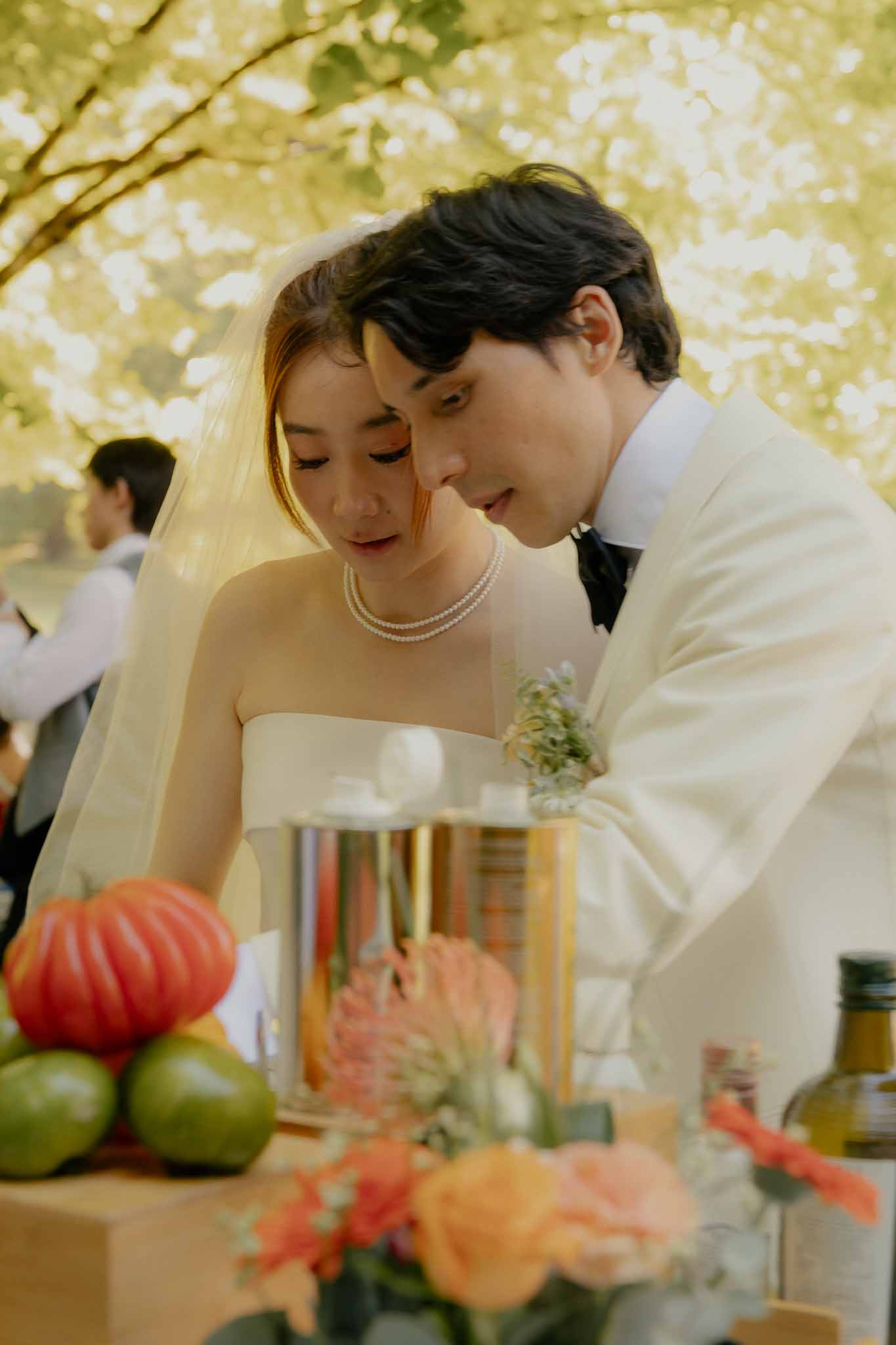 Bride and groom lean over reception table styled with vegetables, coral roses, and champagne in golden light