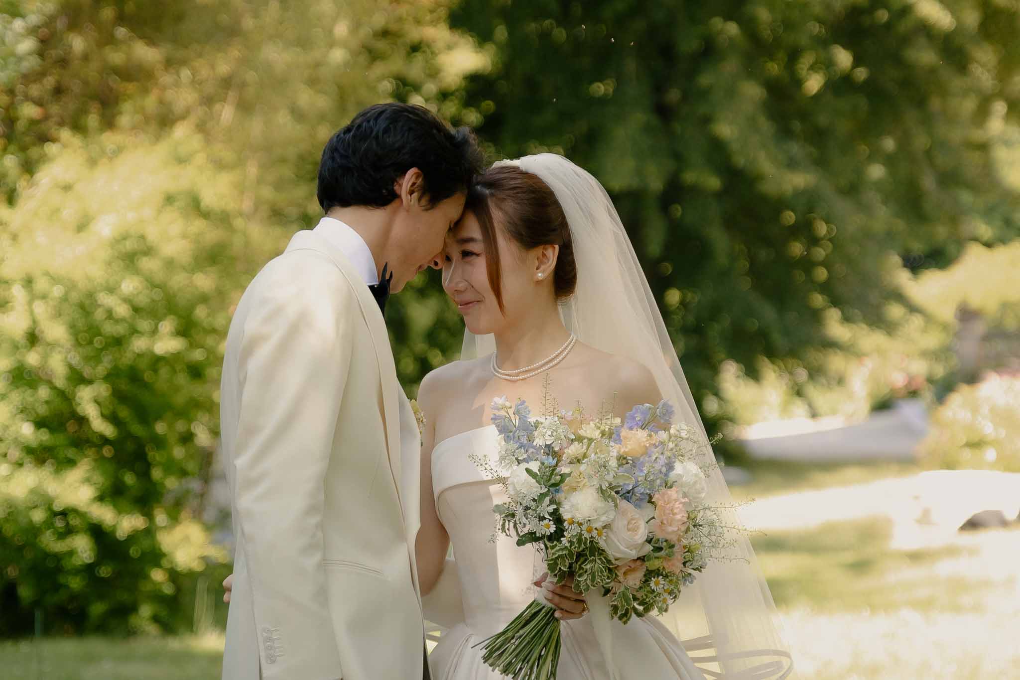 Couple touching foreheads, bride with pearl necklace holding peony and delphinium bouquet in garden