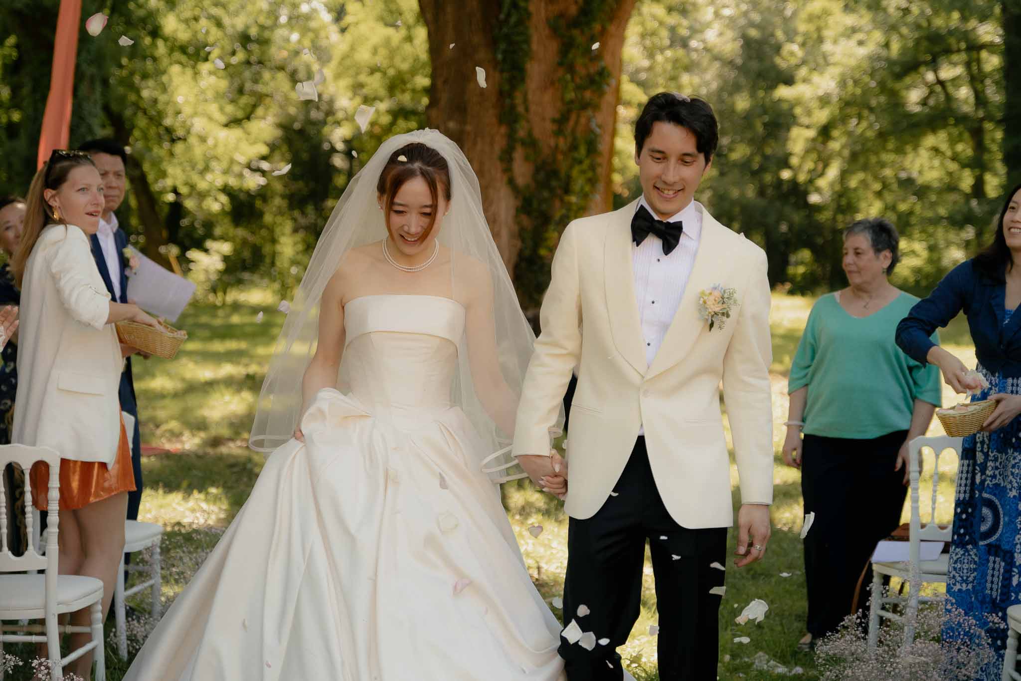 Bride in ivory ballgown and groom in cream dinner jacket walking aisle as guests toss white petals