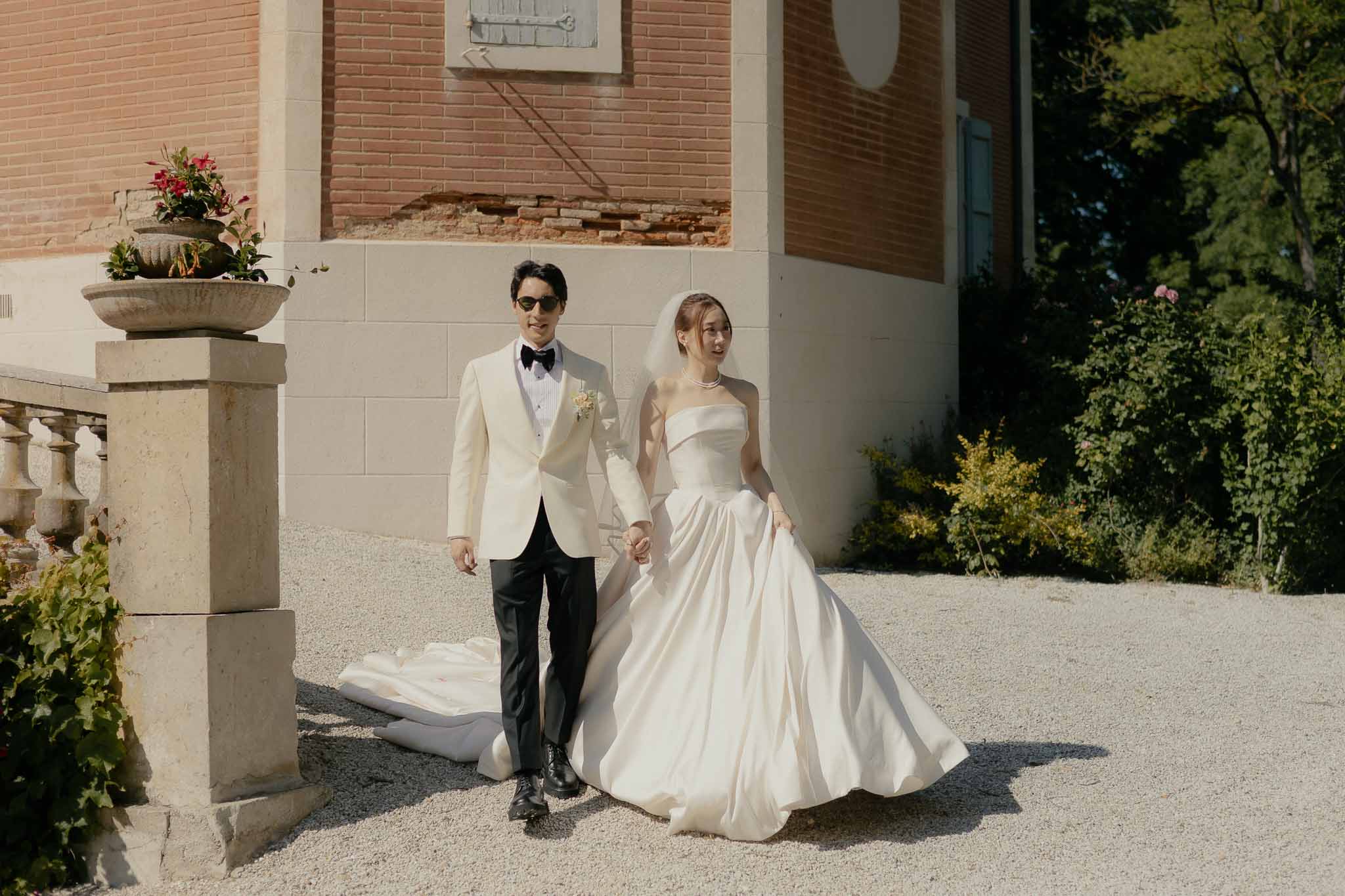 Bride in strapless satin ball gown and groom in cream dinner jacket walking along red-brick chateau path