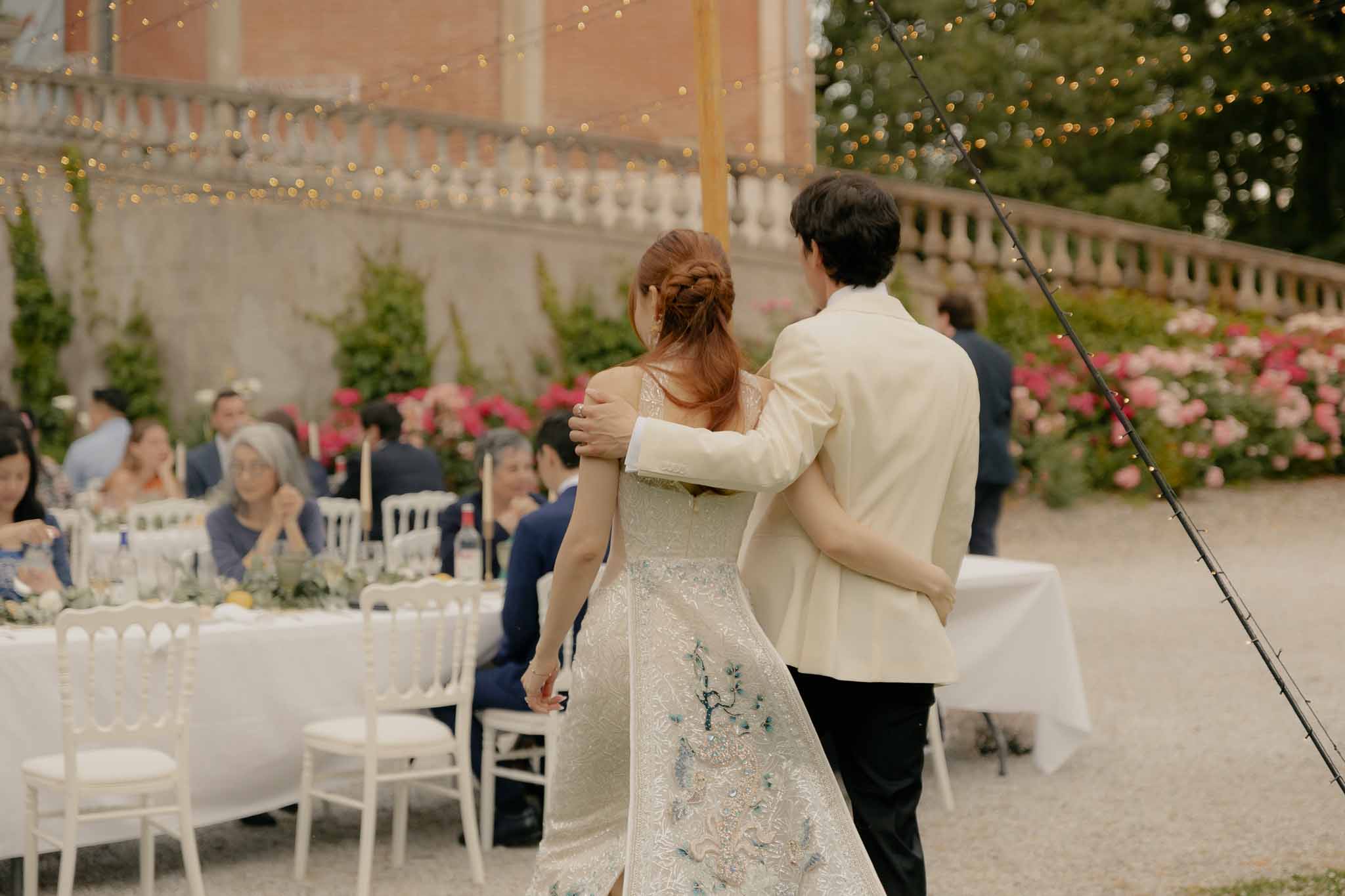 Couple walking arm in arm toward outdoor reception dinner with fairy lights at chateau courtyard