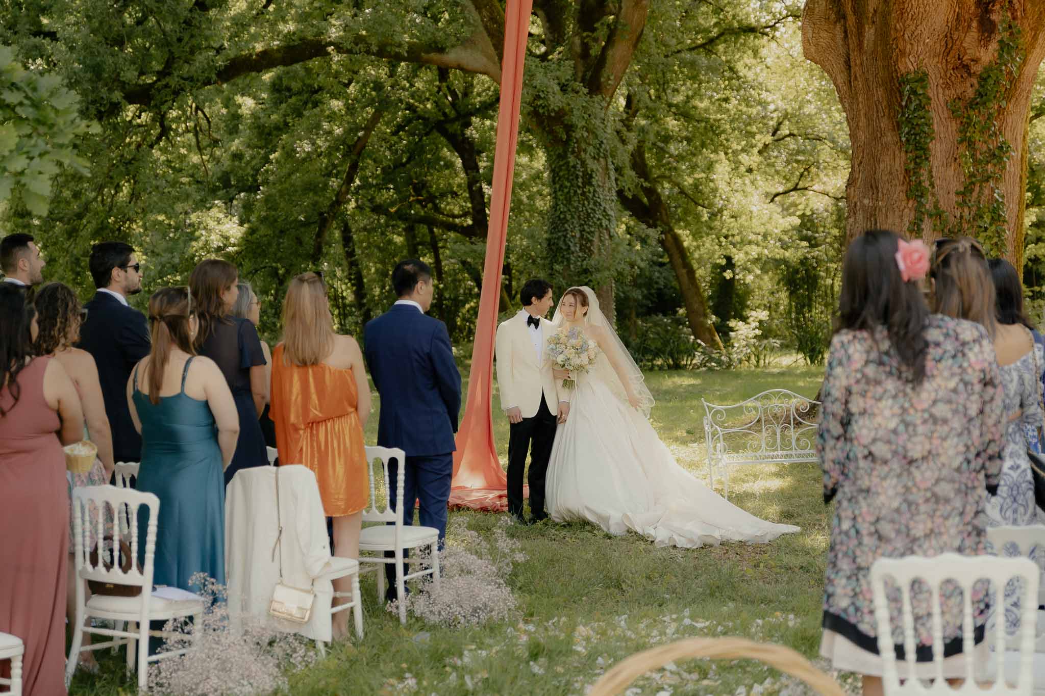 Outdoor woodland ceremony with couple at altar under trees, white Chiavari chairs, and baby's breath aisle