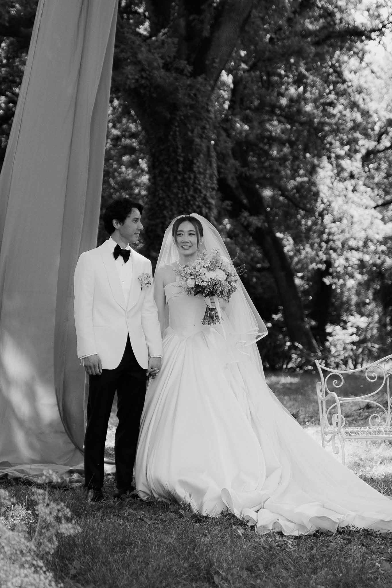 Black and white of couple holding hands in garden, bride in ballgown with veil, groom in white dinner jacket