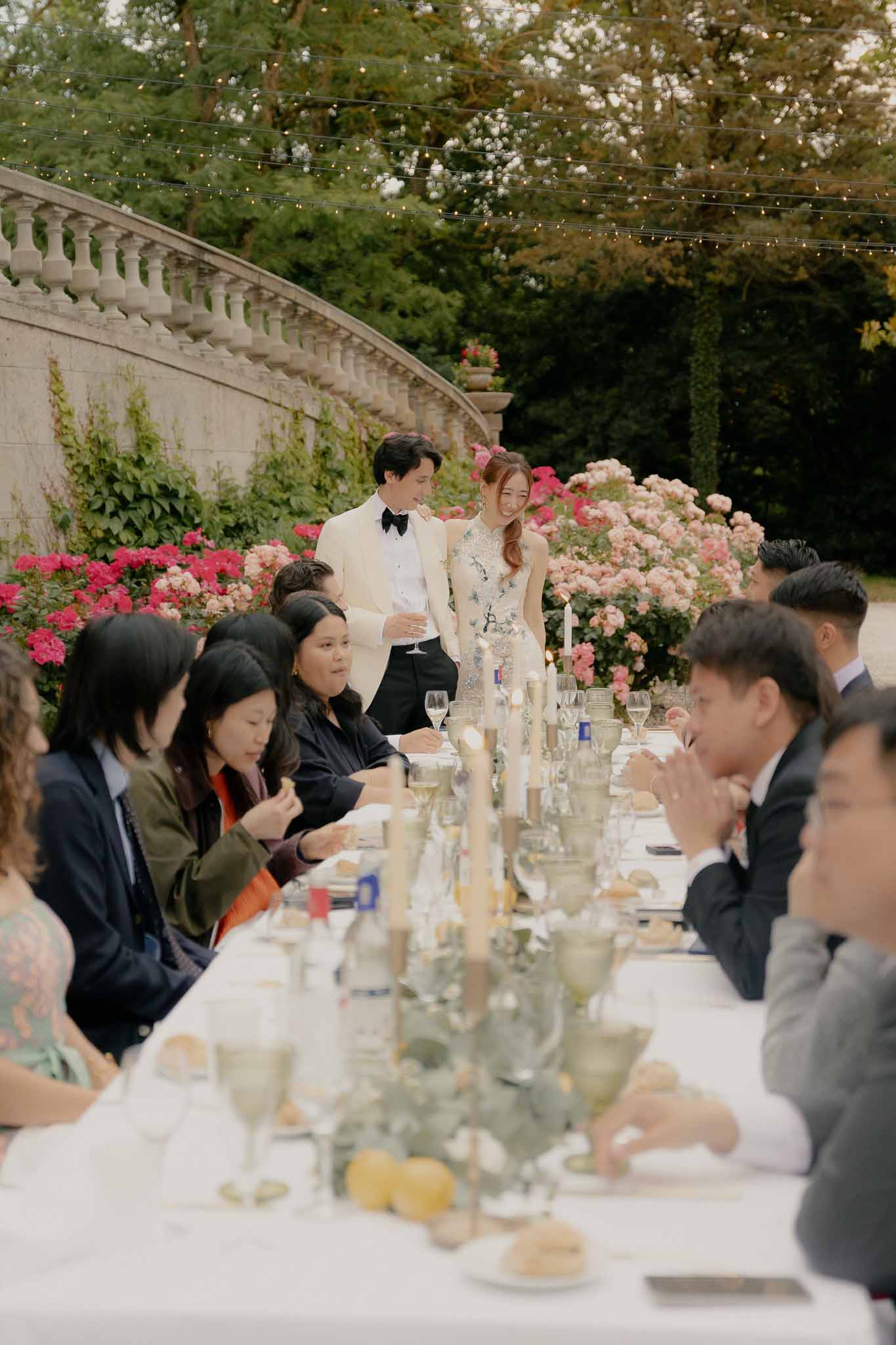 Couple standing at head of banquet table with guests, sage glassware, lemon accents, and fairy lights on garden terrace