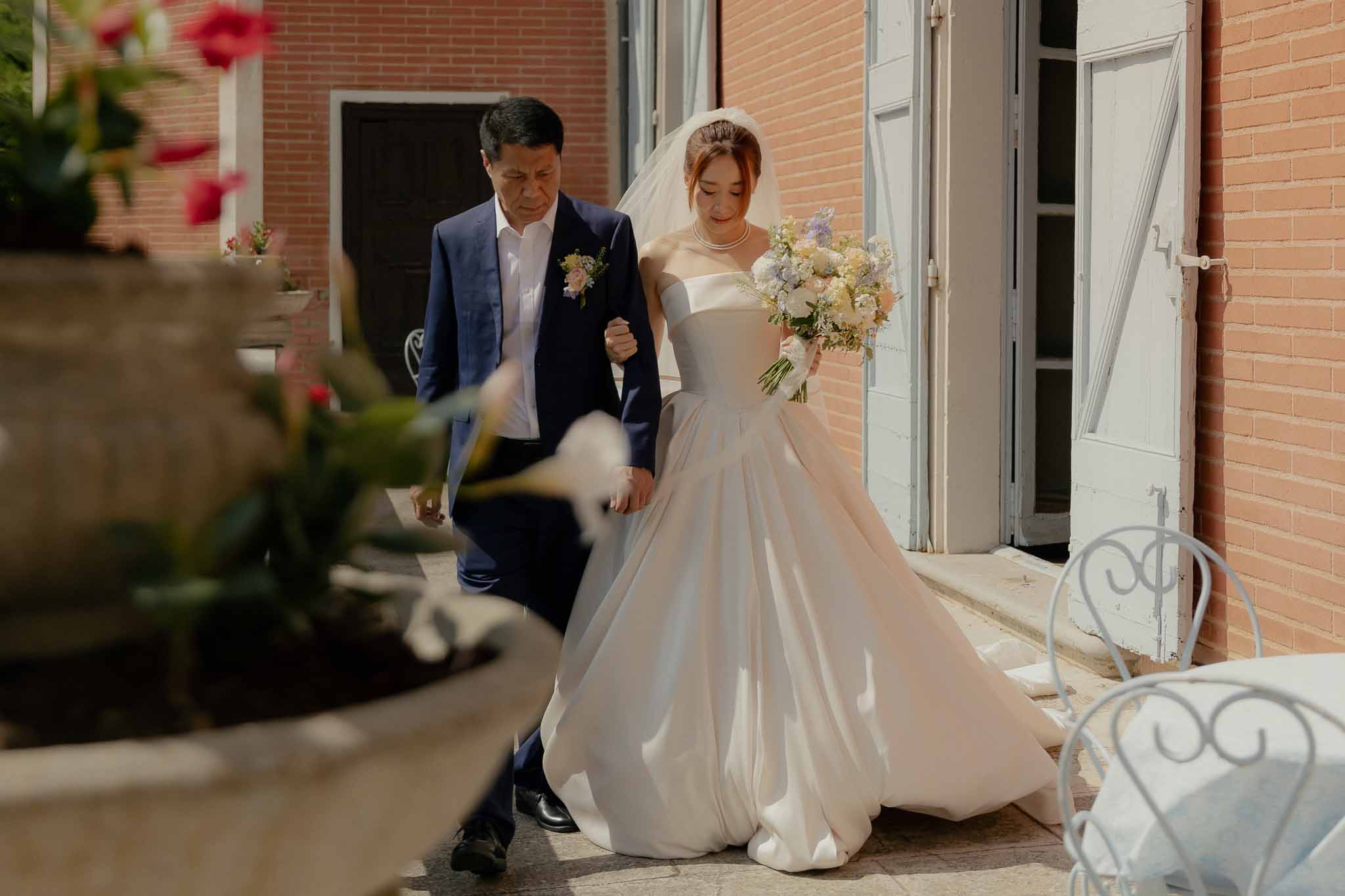 Father escorting bride in ivory ballgown with lavender and blush bouquet along red brick terrace