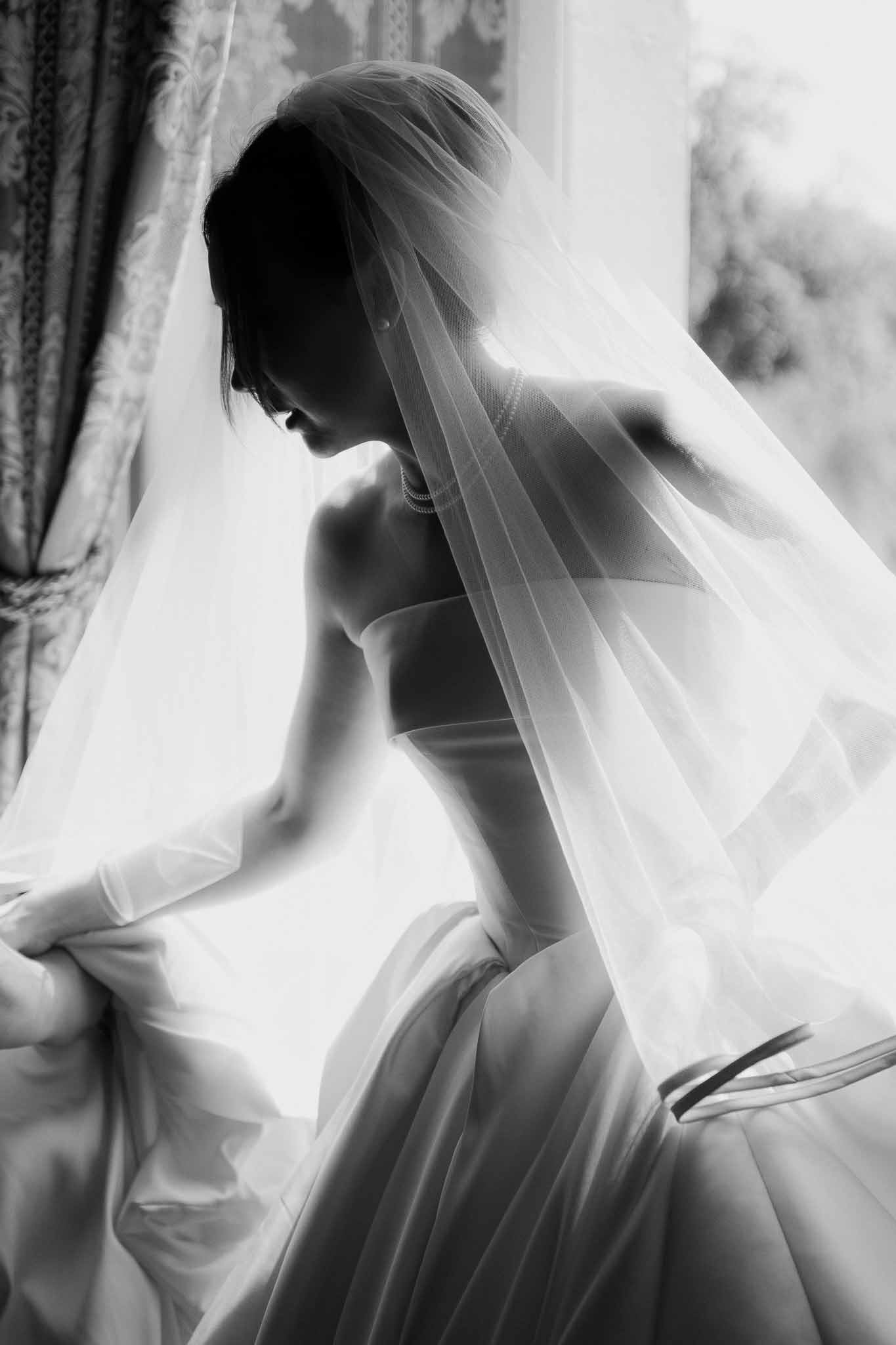 Black and white silhouette of bride in strapless gown adjusting veil by bright window with pearl necklace