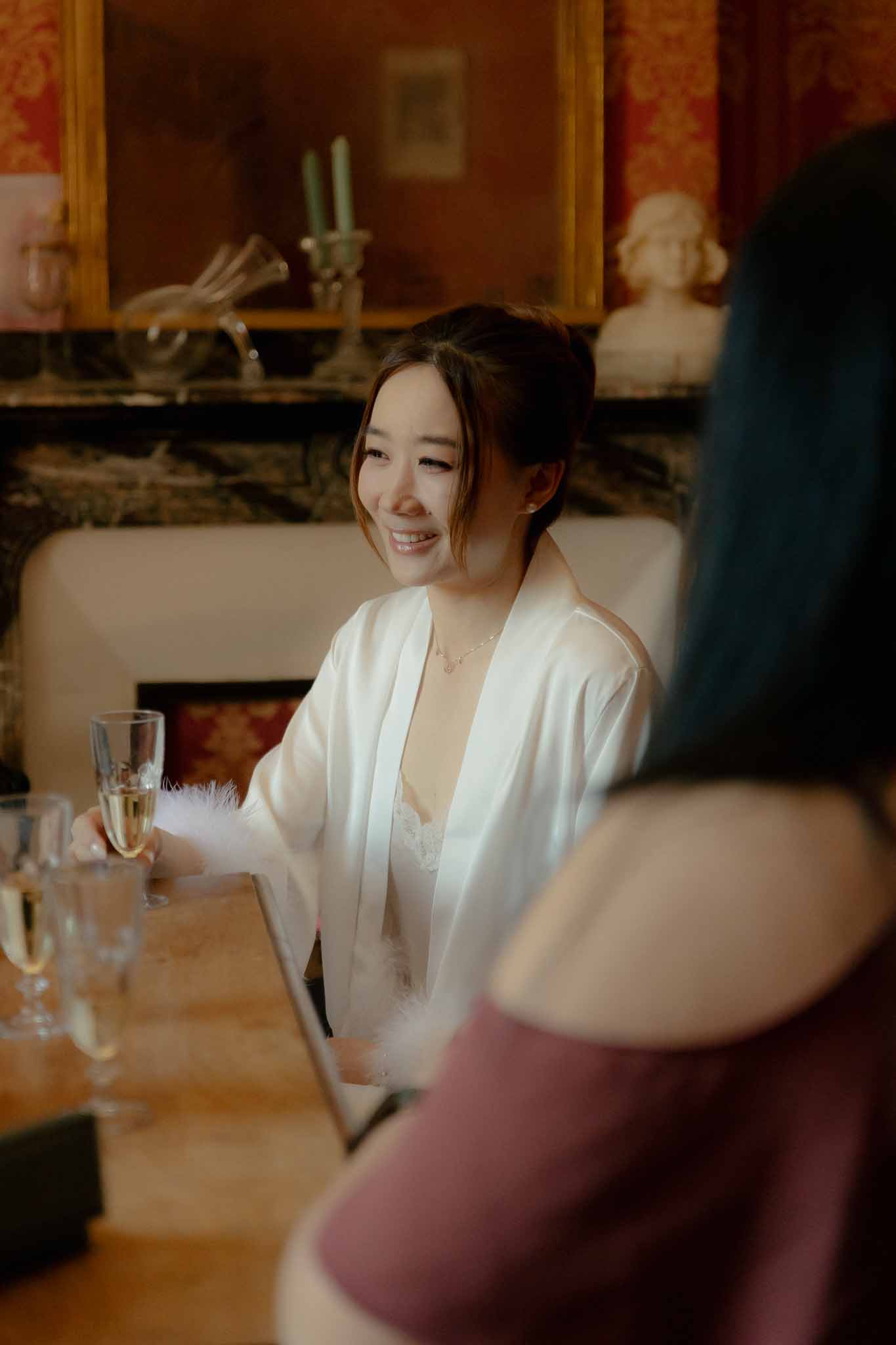 Bride in feather-trimmed robe holds champagne by green marble fireplace in red damask chateau salon