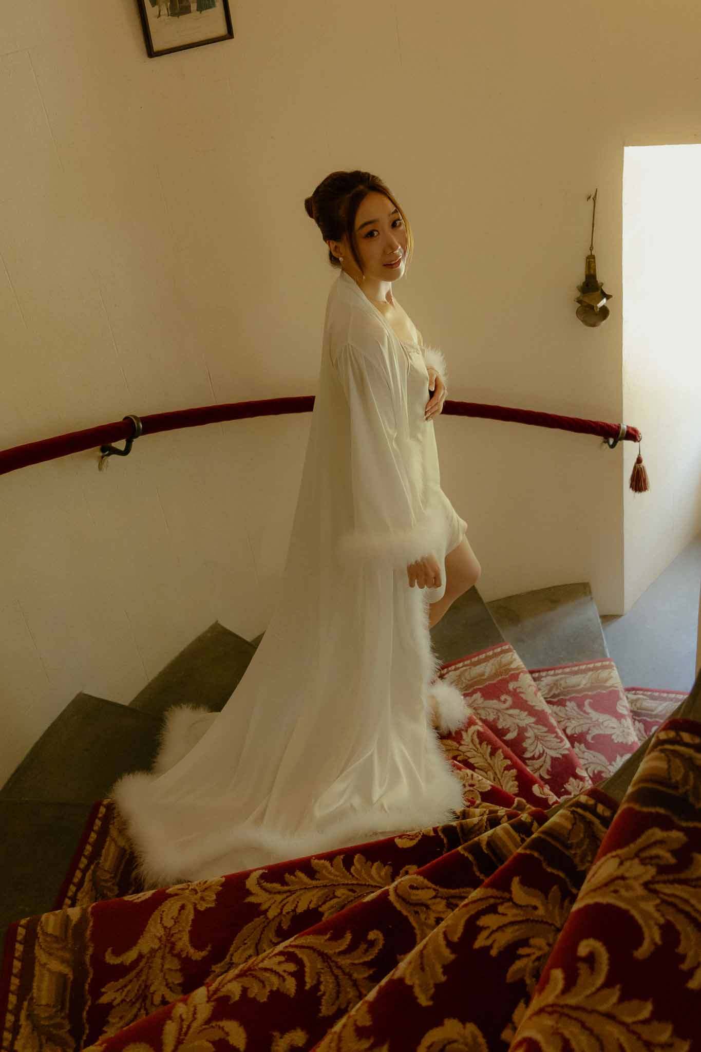 Bride in white feather-trimmed robe looking back on crimson velvet staircase with gold damask