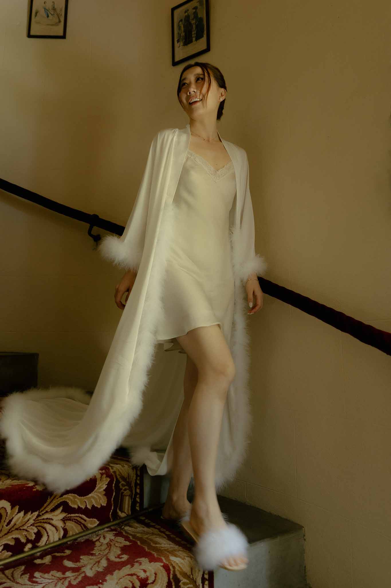 Bride laughing on staircase in white feather-trimmed robe and matching fluffy slippers