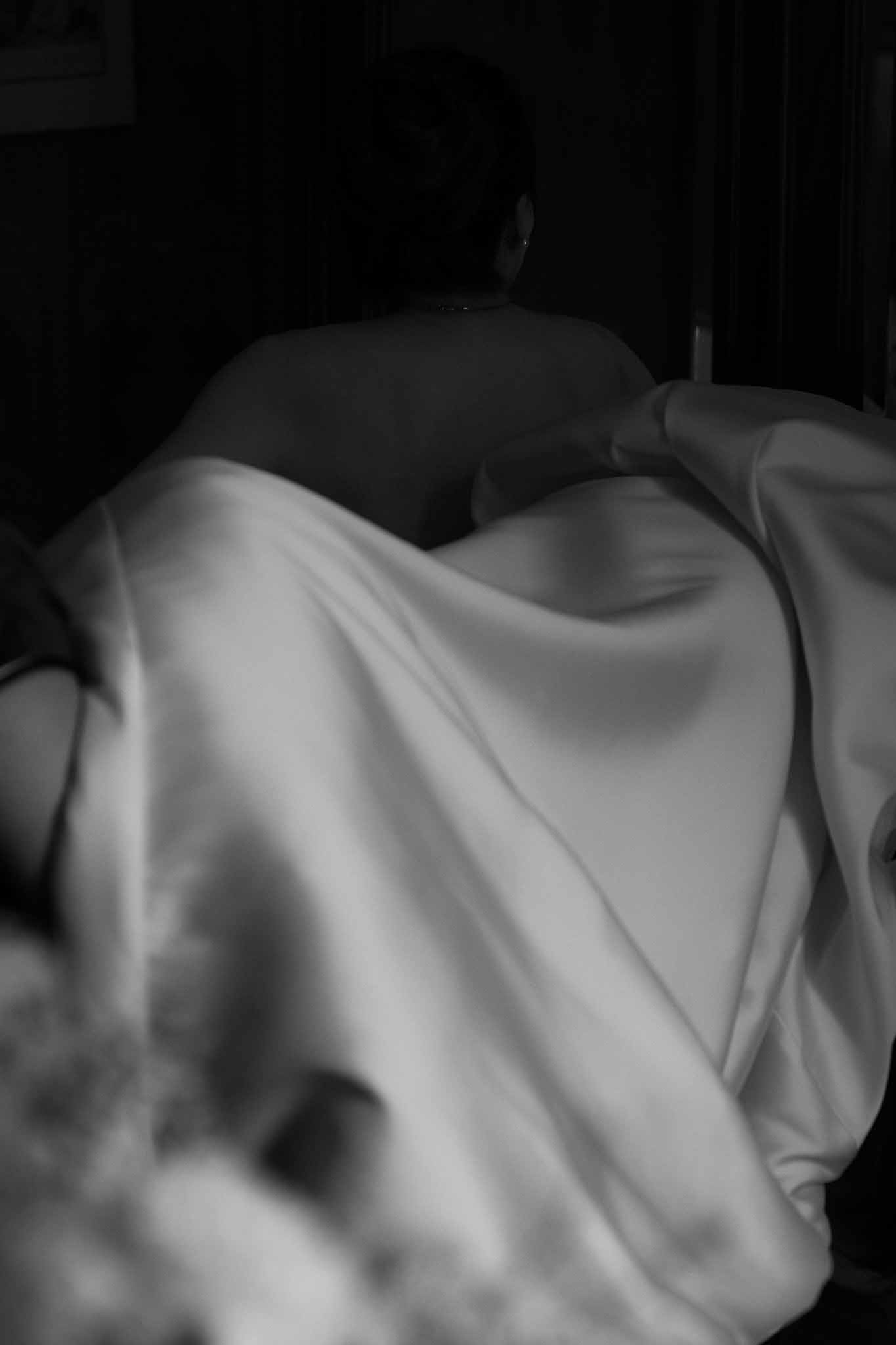 Black and white image of bridesmaid holding up back of bride's satin wedding dress in dark room