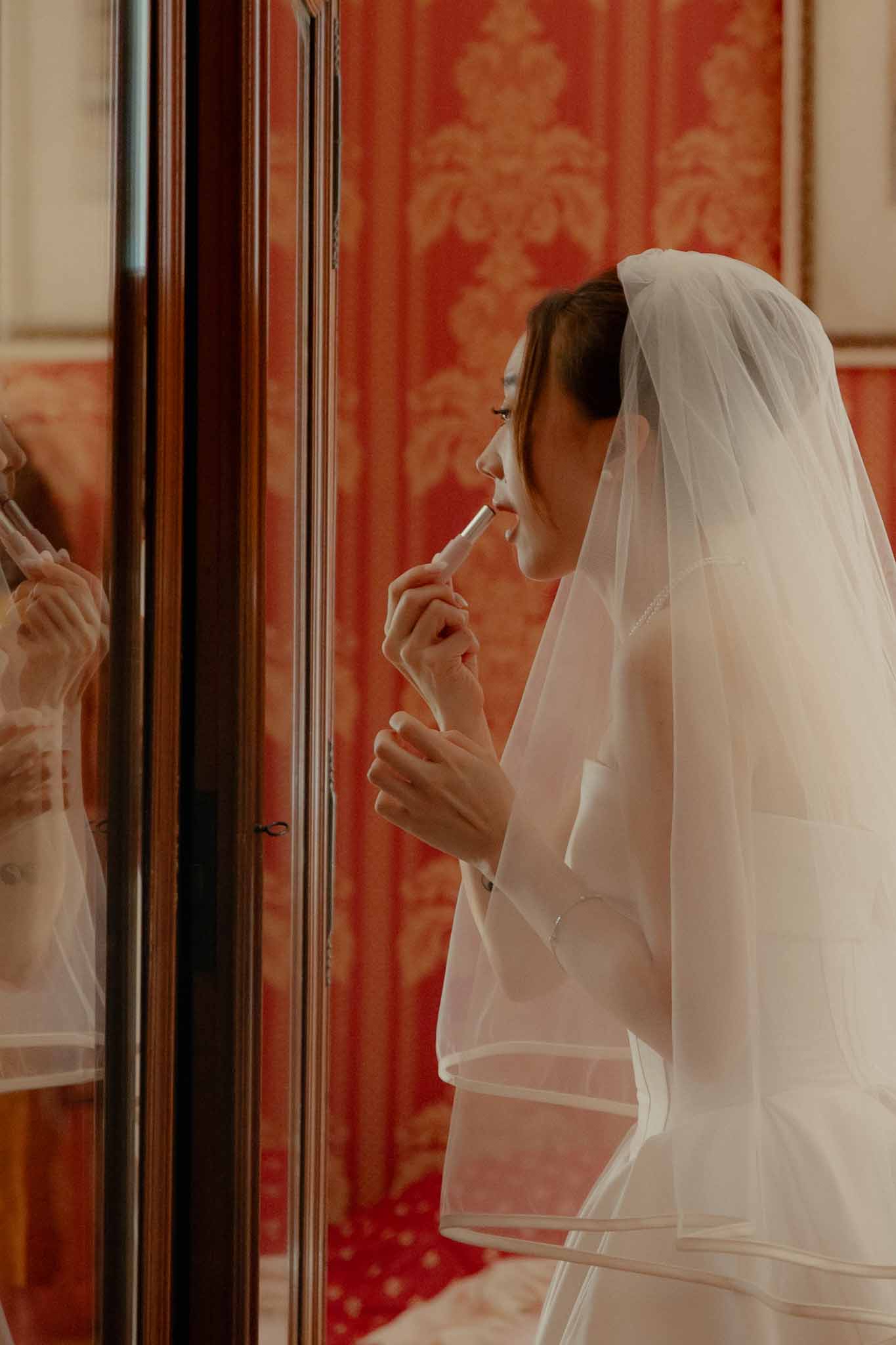 Bride in white gown and tulle veil applying lipstick at mirror against red damask wallpaper
