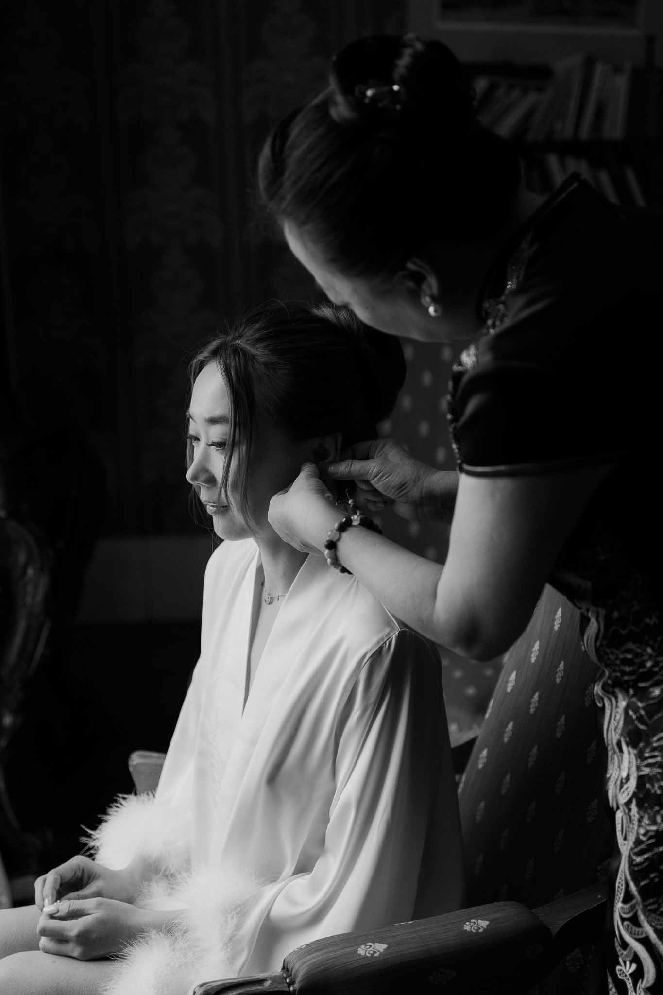 Black and white bride in feather-trimmed robe having updo styled by attendant in dark interior room