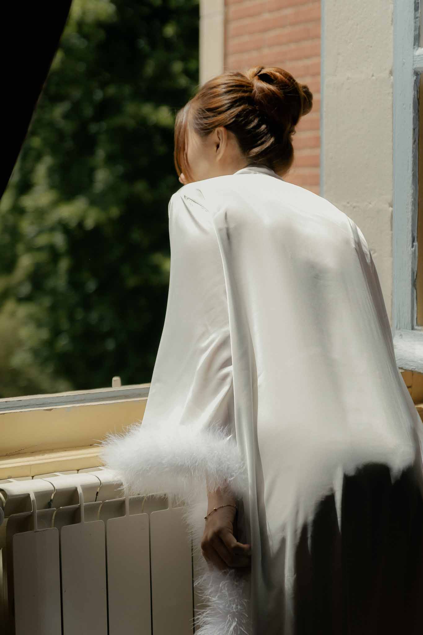 Bride in white satin robe with feather-trimmed sleeves looking out window during getting ready with hair in updo
