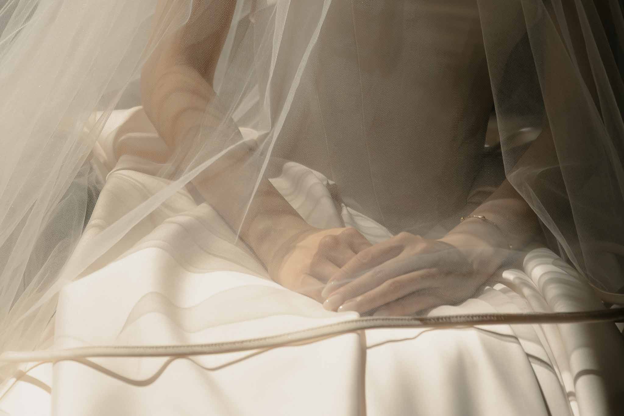 Close-up of couple's hands beneath flowing ivory tulle veil with warm diffused light