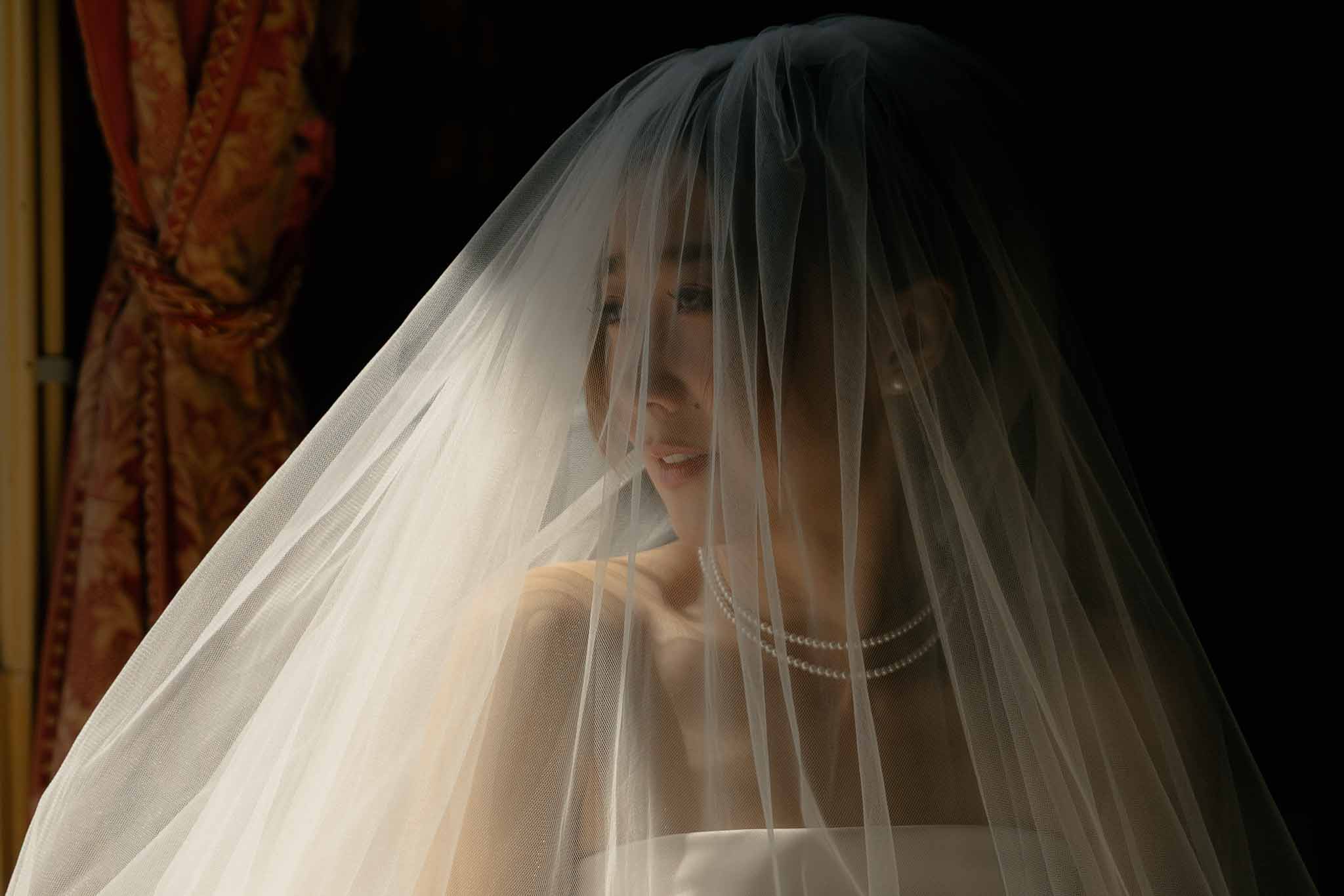 Bride with tulle veil draped over face and pearl necklace in dramatic chiaroscuro lighting