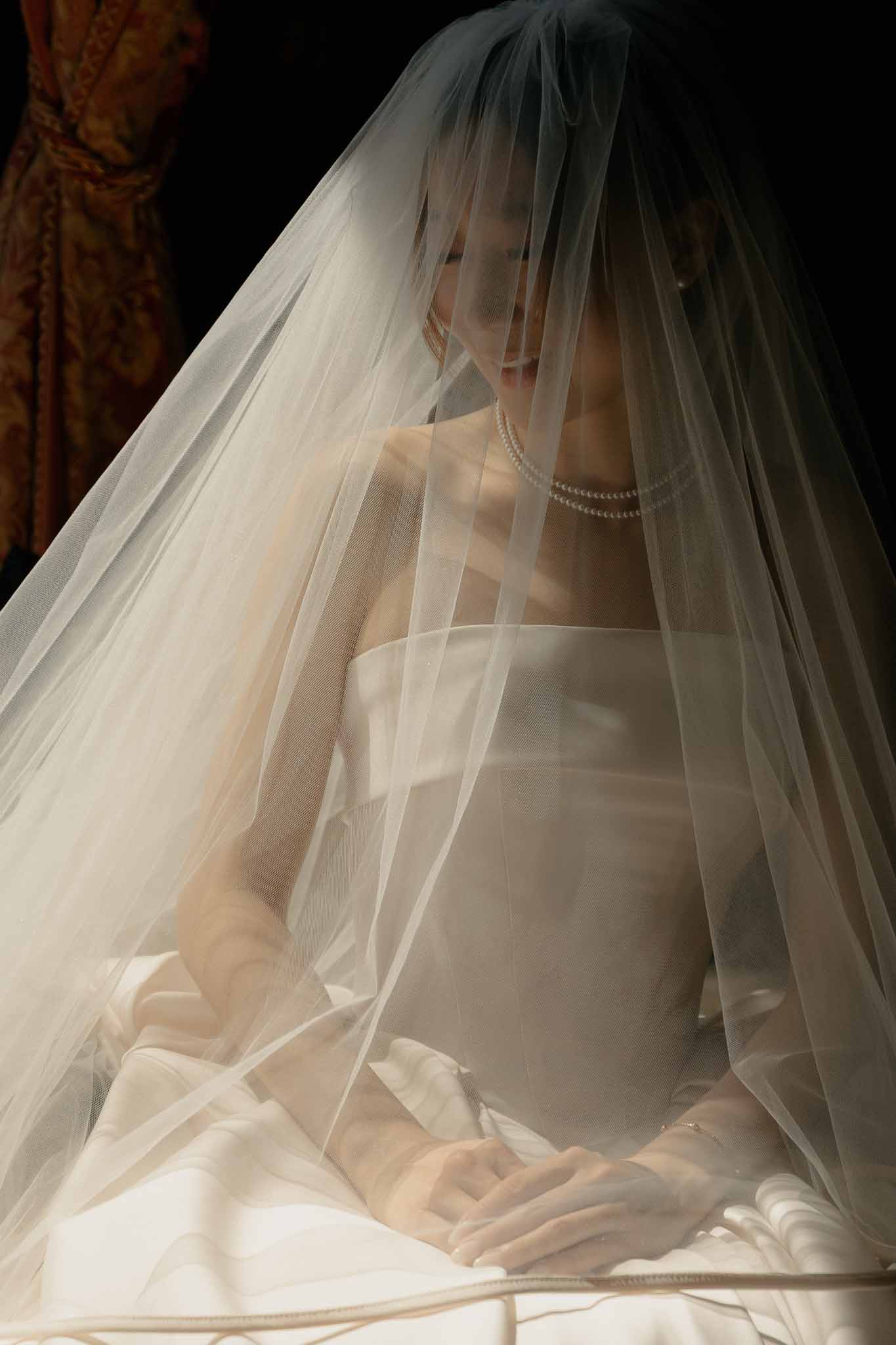 Bride seated indoors draped in voluminous ivory tulle veil with pearl necklace and dark background