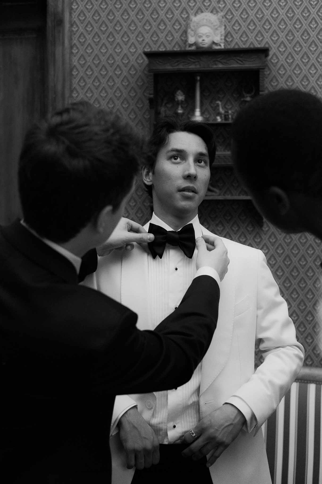 Black and white groomsmen adjusting grooms bow tie in white dinner jacket in ornate chateau room with patterned wallpaper