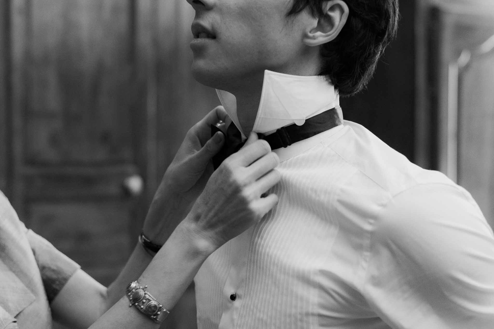 Black and white close-up of bow tie being fastened on groom's pleated tuxedo shirt