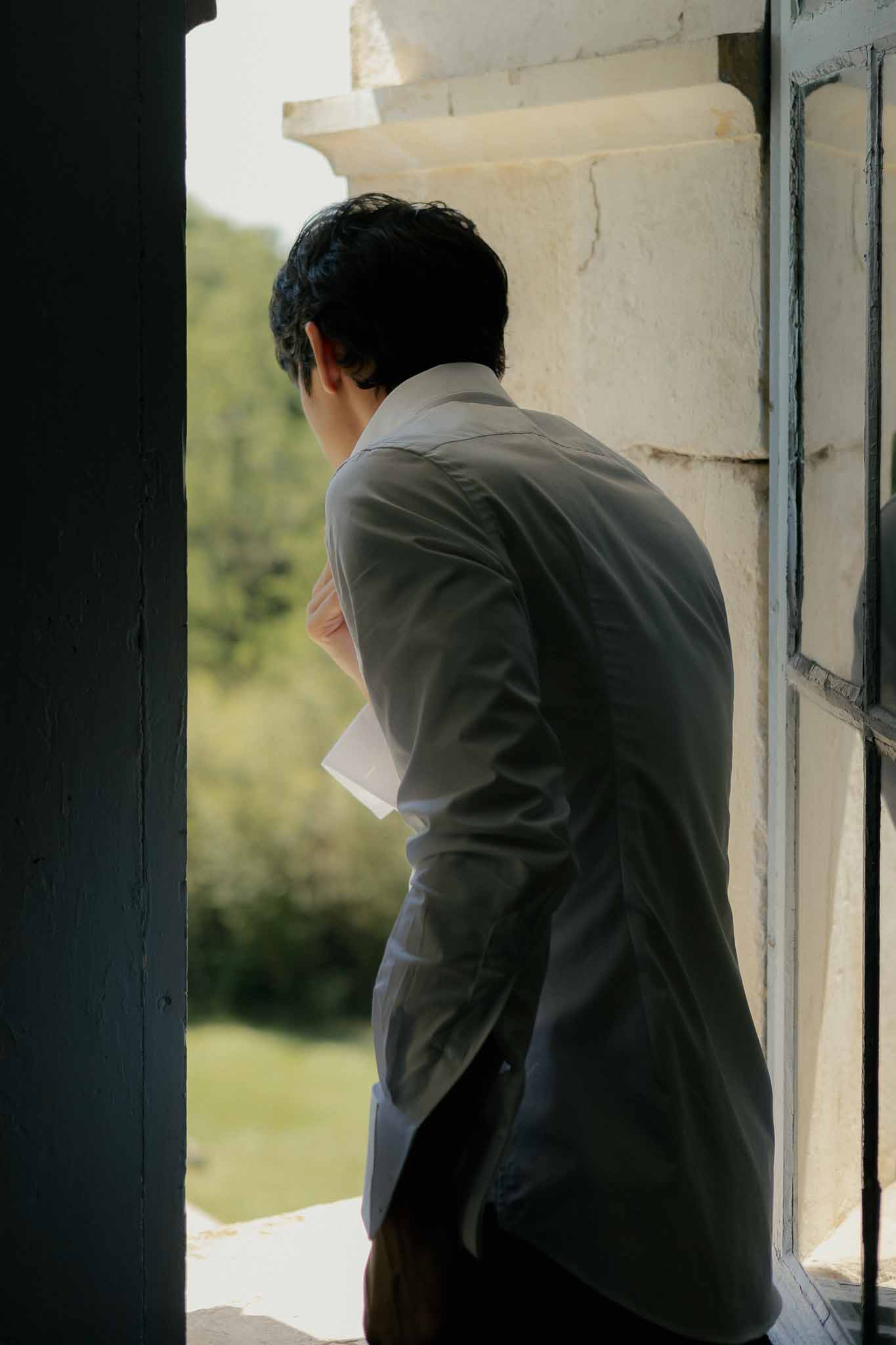 Groom in grey suit reading a letter from his bride in a stone building doorway before the ceremony