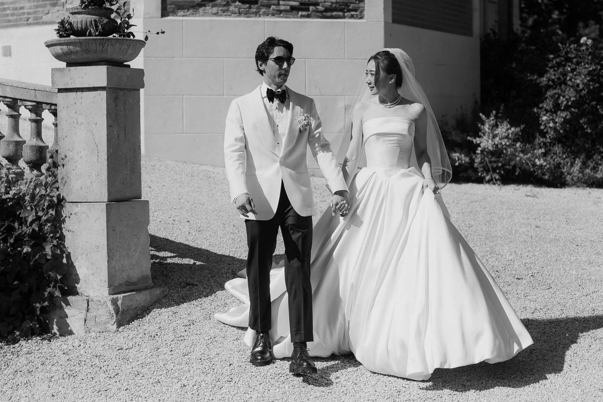 Black and white portrait of bride in strapless ballgown and groom in white dinner jacket walking on chateau gravel courtyard