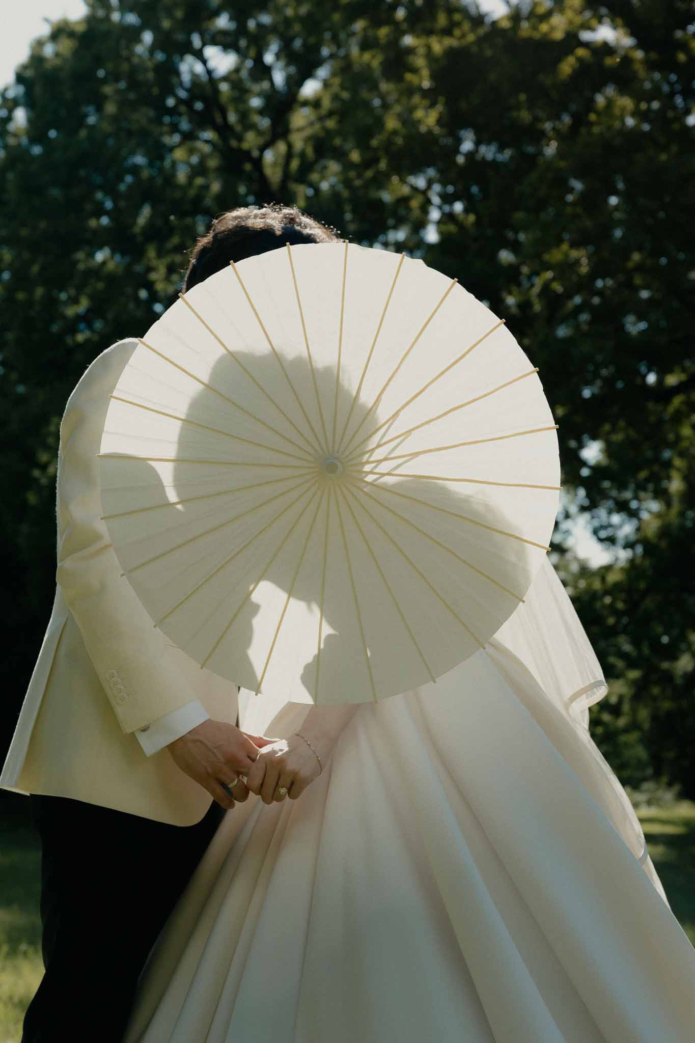 Couple holding an ivory paper parasol that obscures their faces, with veil trailing behind and wedding rings visible