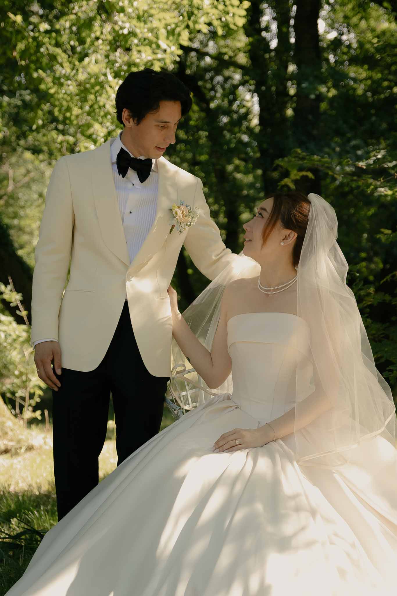 Bride in ivory ballgown with pearl necklace and cathedral veil seated beside groom in cream dinner jacket in garden