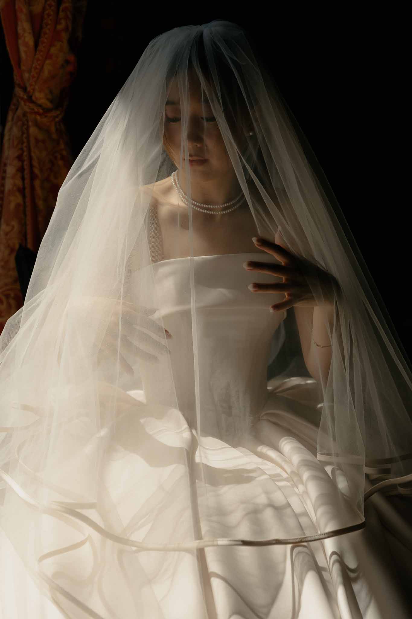 Bride seated with veil draped over her head wearing strapless ivory satin gown and pearl necklace in moody light