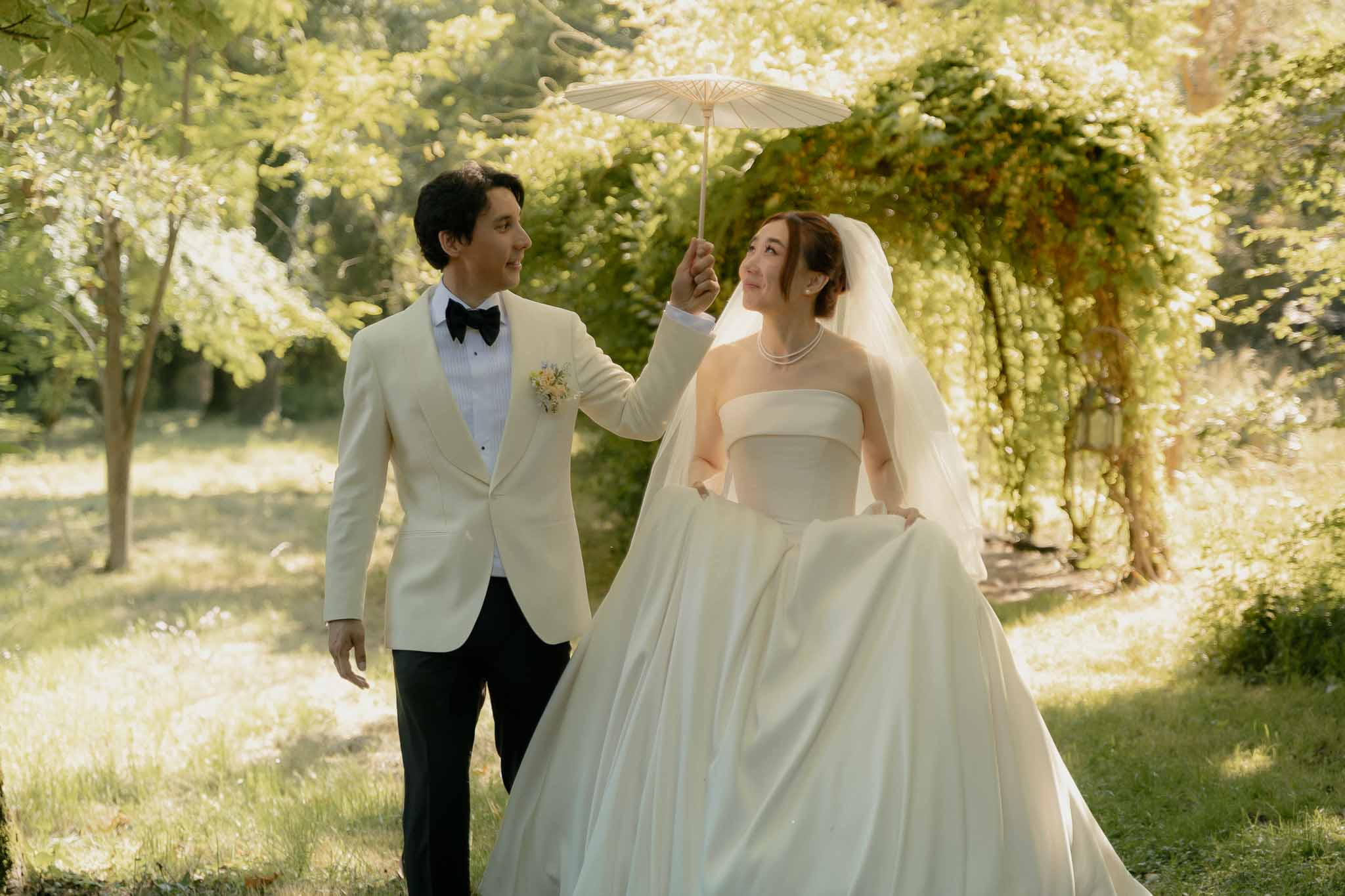 Groom in cream dinner jacket and bride with pearl necklace and paper parasol walking in sunlit garden