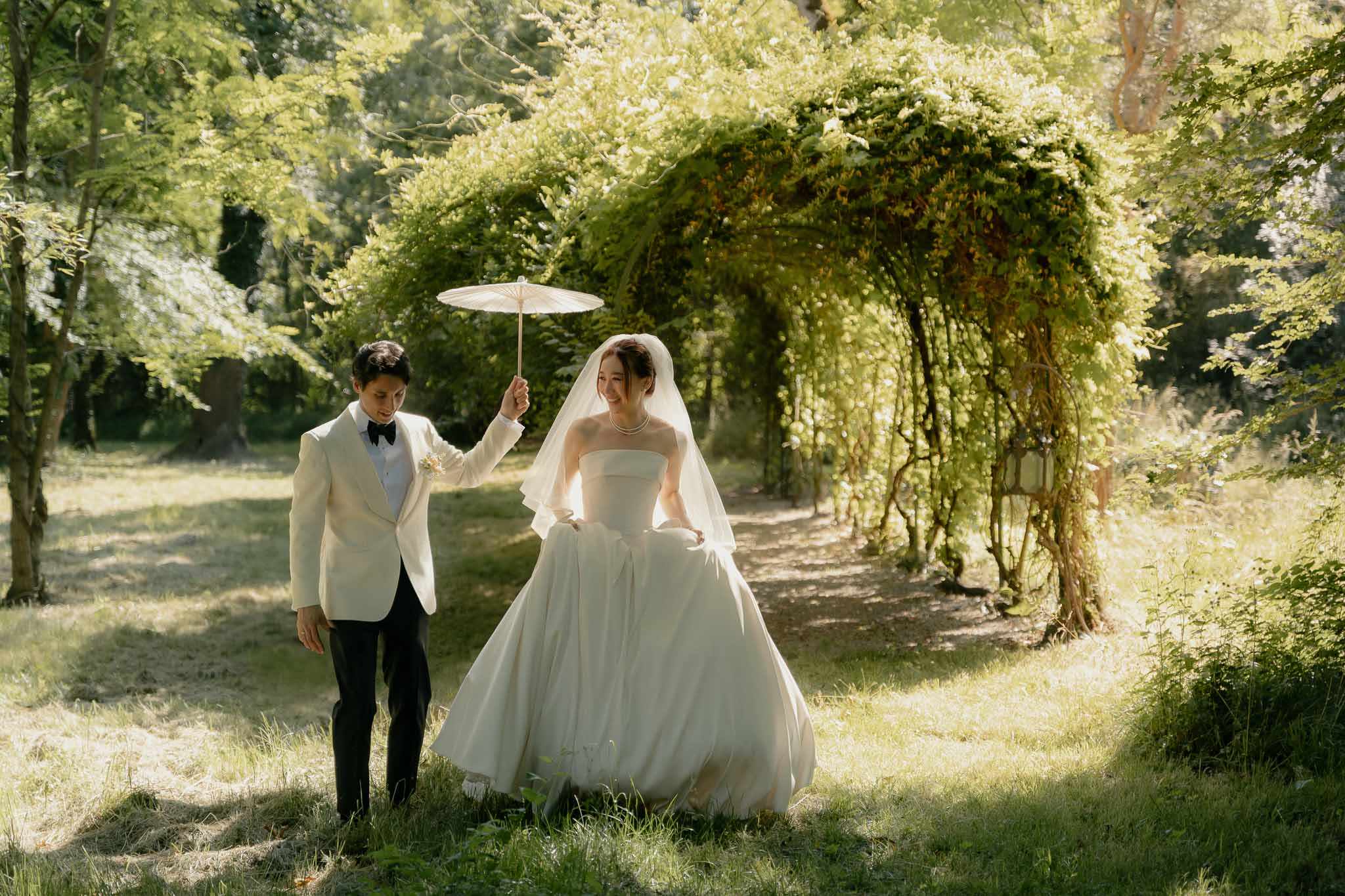 Bride and groom walking through a sculpted green garden archway, groom holding a cream parasol over the bride
