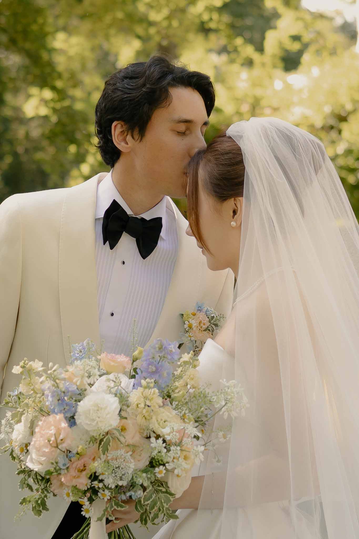 Groom in ivory tuxedo kissing bride's forehead as she holds pastel garden-style bouquet
