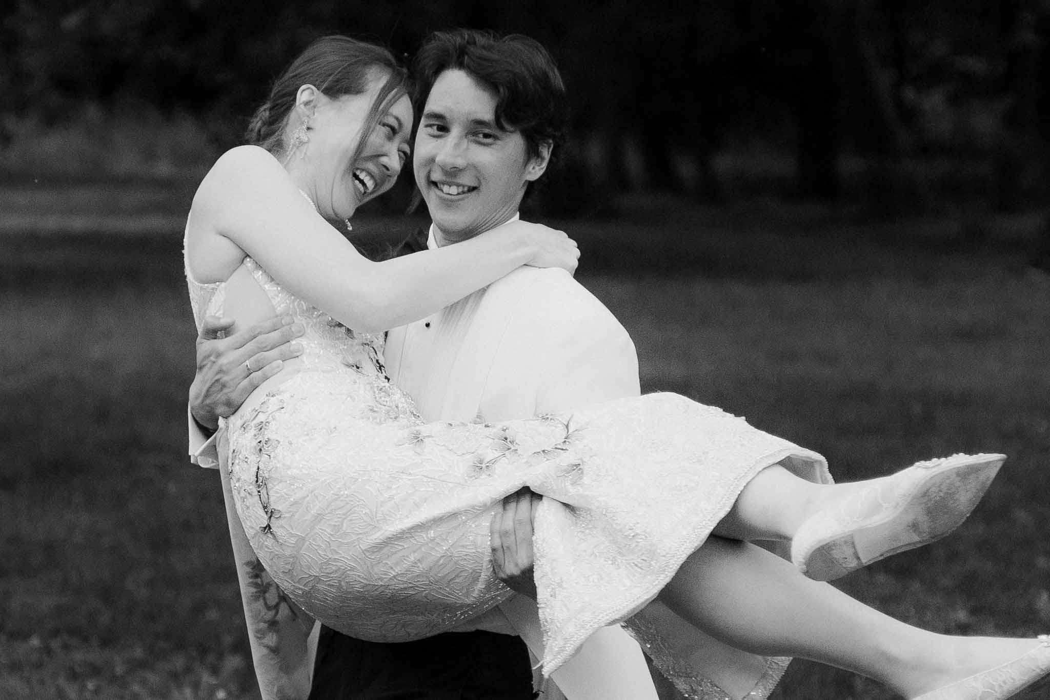 Black-and-white portrait of groom carrying laughing bride in his arms on a lawn with trees behind