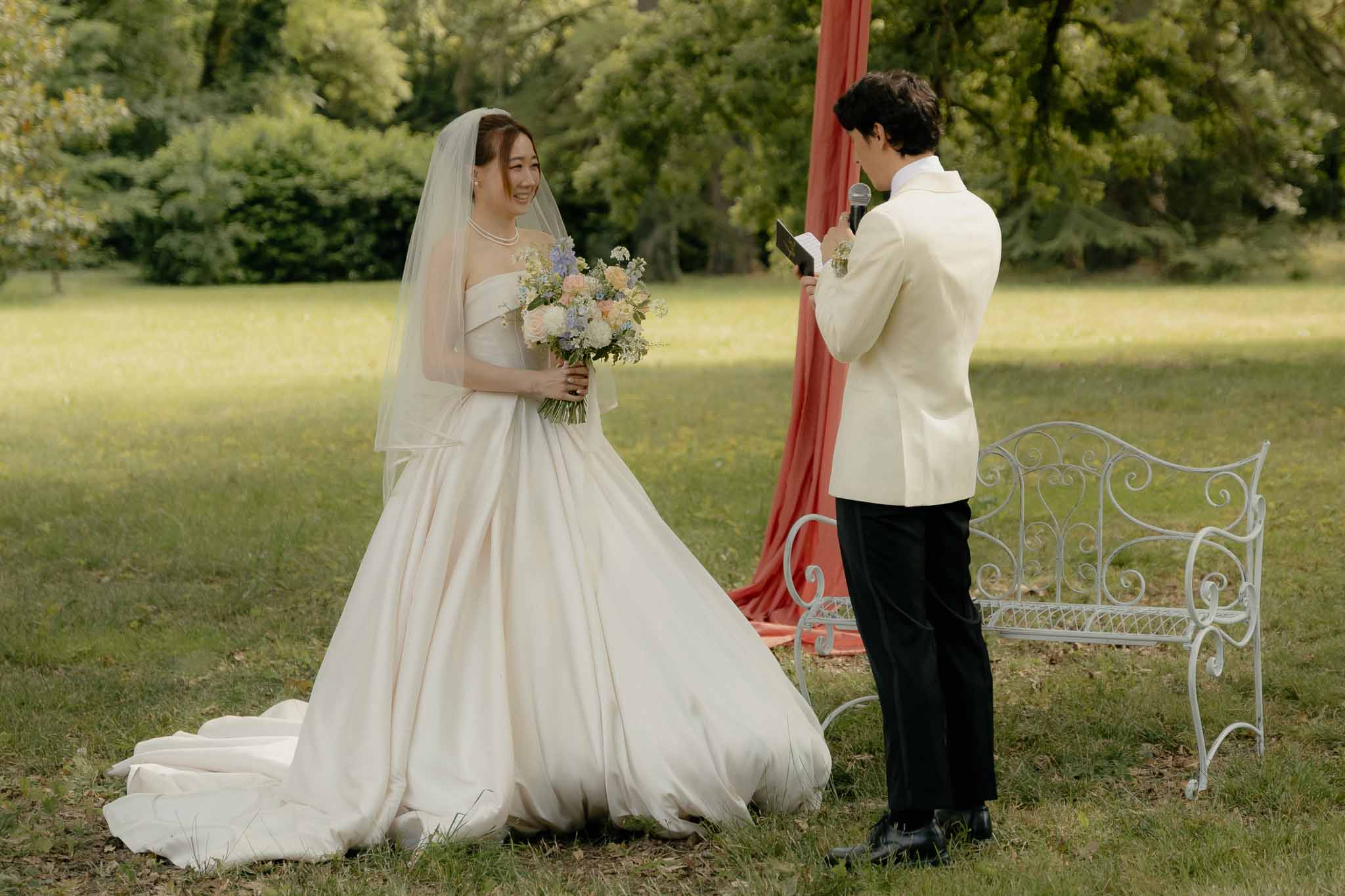 Groom reading vows in cream jacket as bride holds peach rose and blue hydrangea bouquet on garden lawn