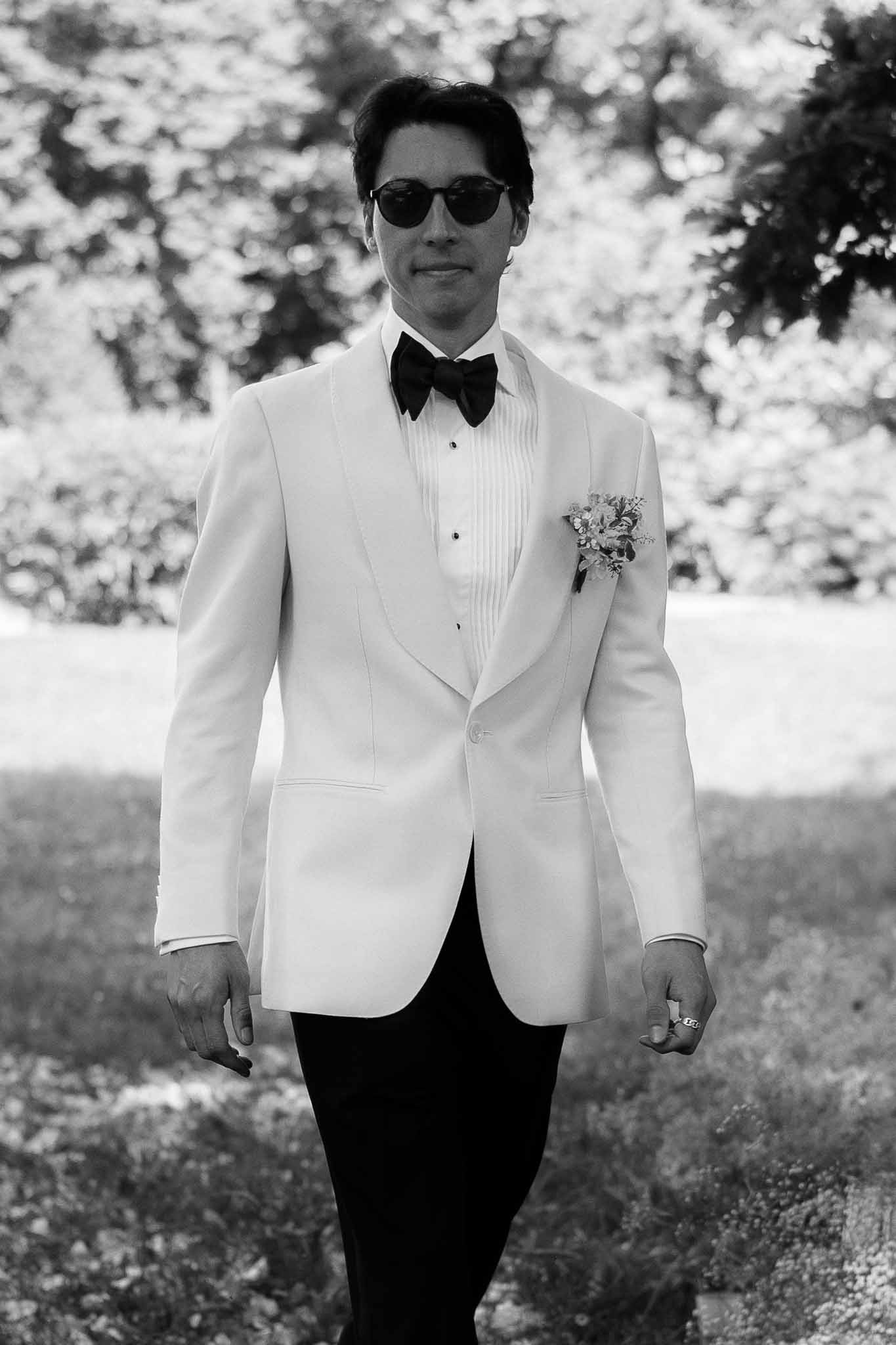 Black and white portrait of groom in white dinner jacket with round sunglasses in garden