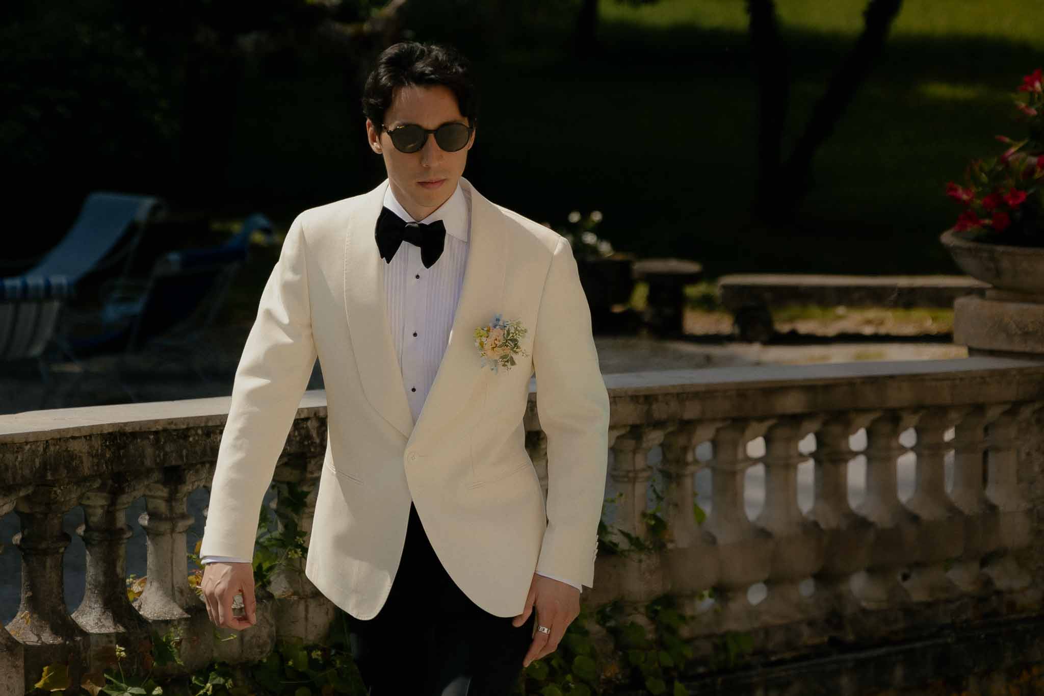 Groom in cream dinner jacket with black velvet bow tie and peach boutonniere on terrace