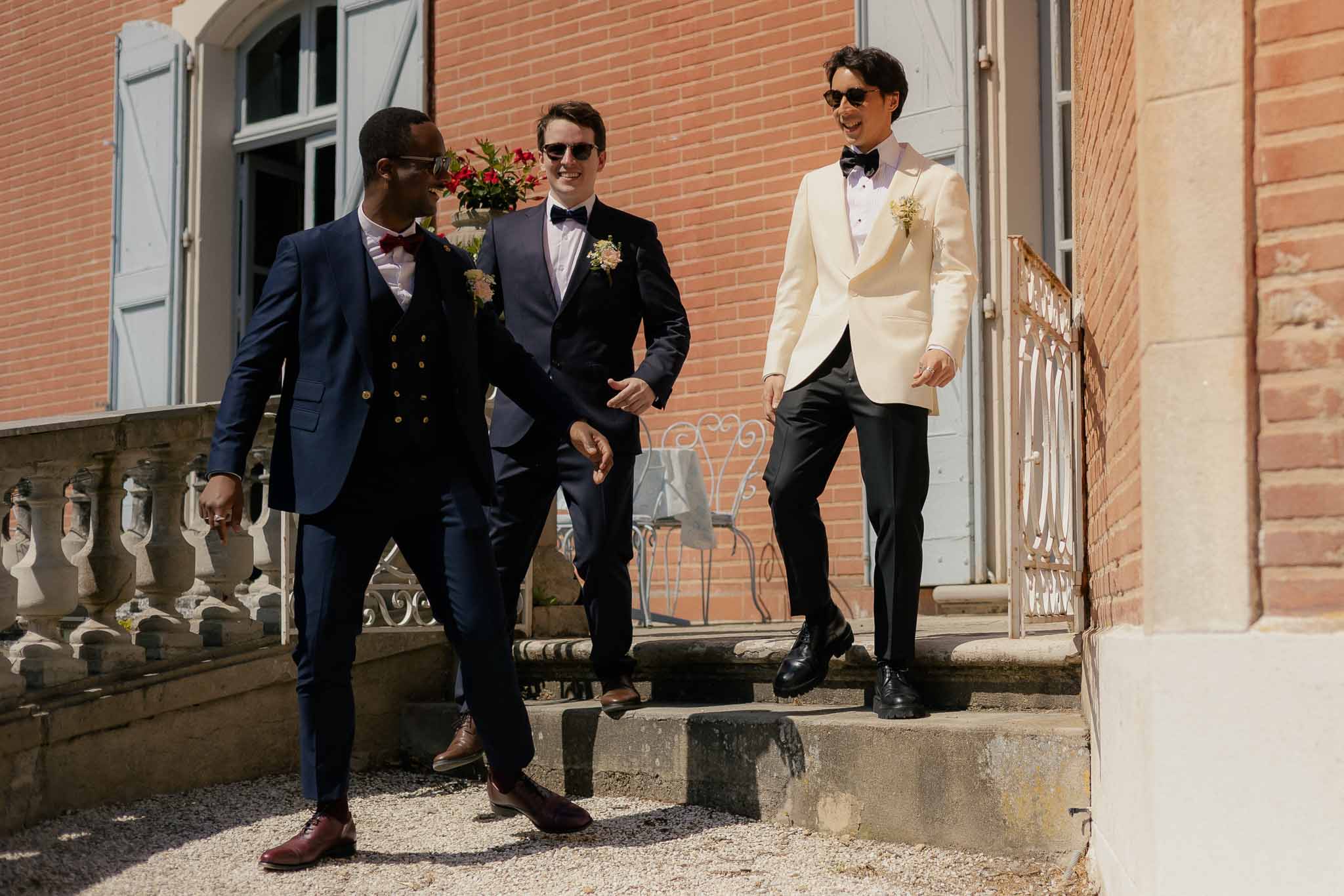 Groom in ivory dinner jacket and two groomsmen in navy suits walking down chateau stone steps laughing
