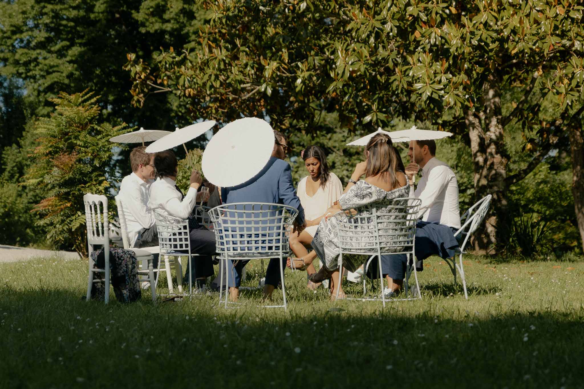 Wedding guests seated with white parasols during outdoor cocktail hour on garden lawn