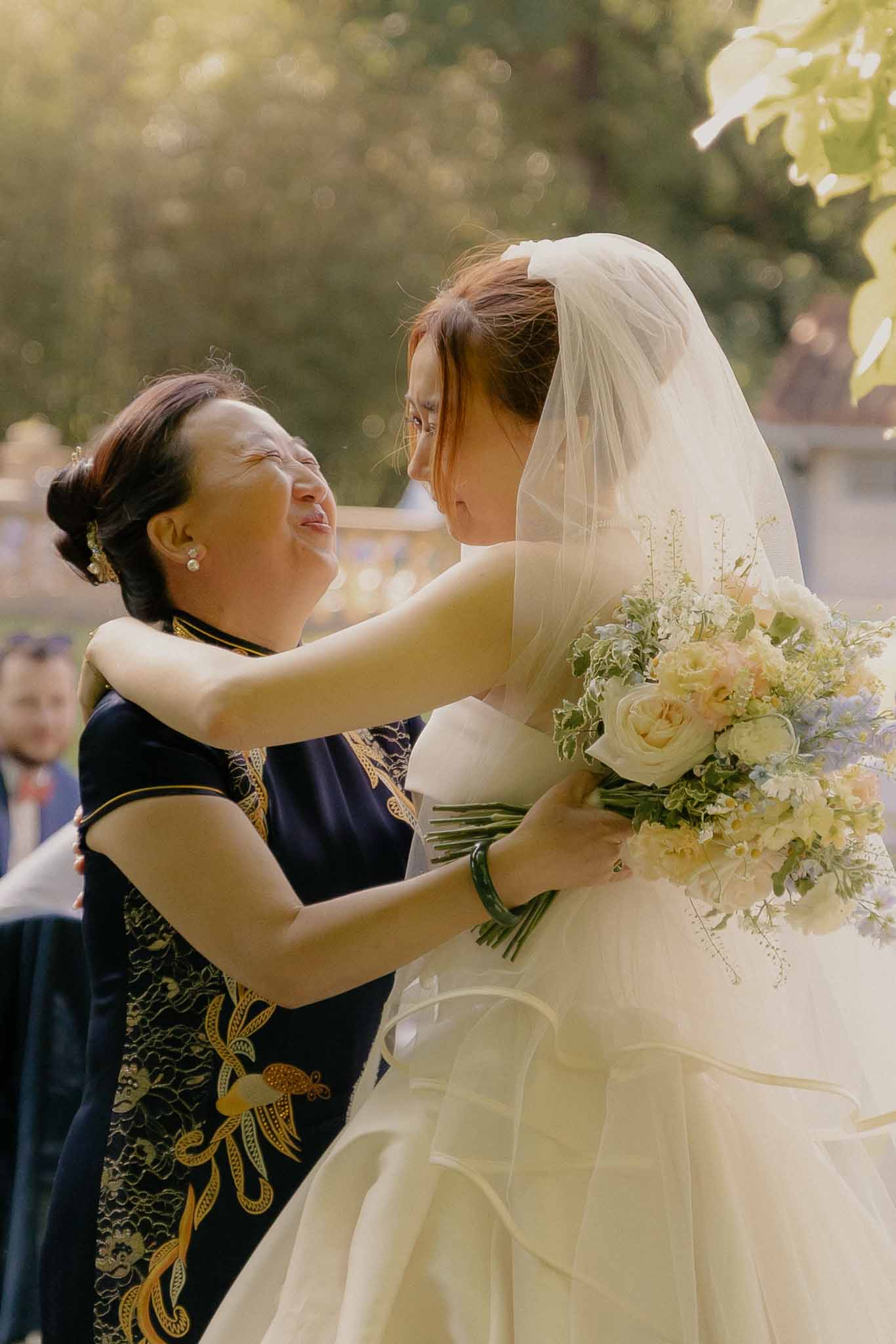 Emotional bride and mother embracing during golden hour, bride holding garden-style bouquet