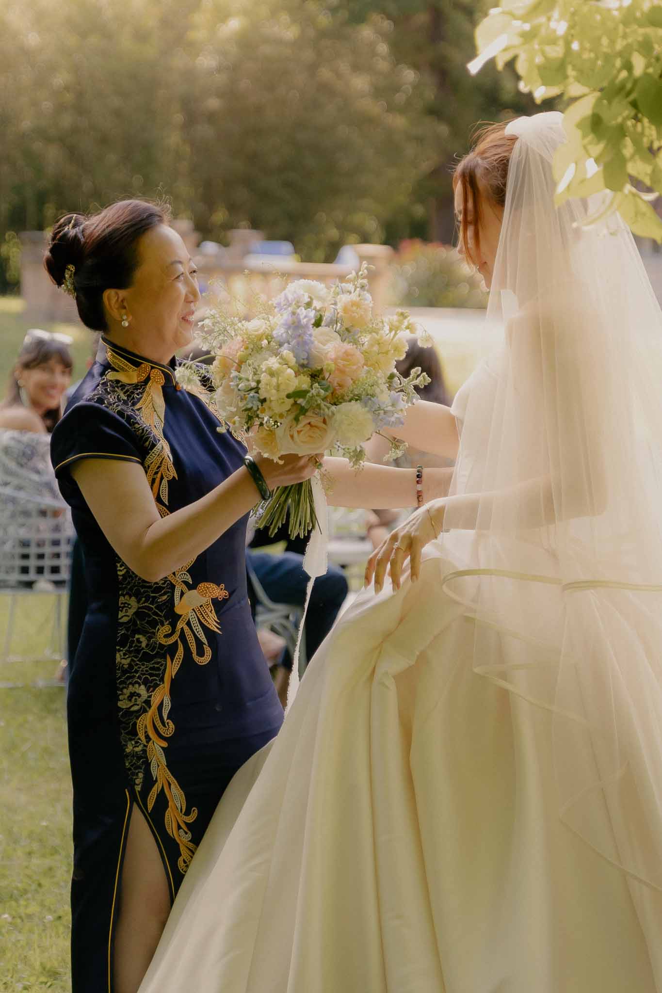 Mother in navy embroidered qipao presenting garden-style bouquet to bride in golden light