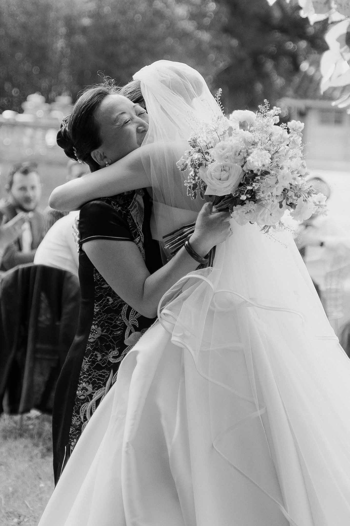 Black and white image of bride embracing older woman after ceremony with guests in background