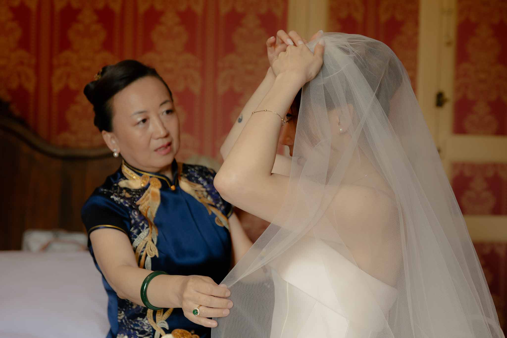 Woman in navy silk qipao placing tulle veil on bride in chateau getting-ready room