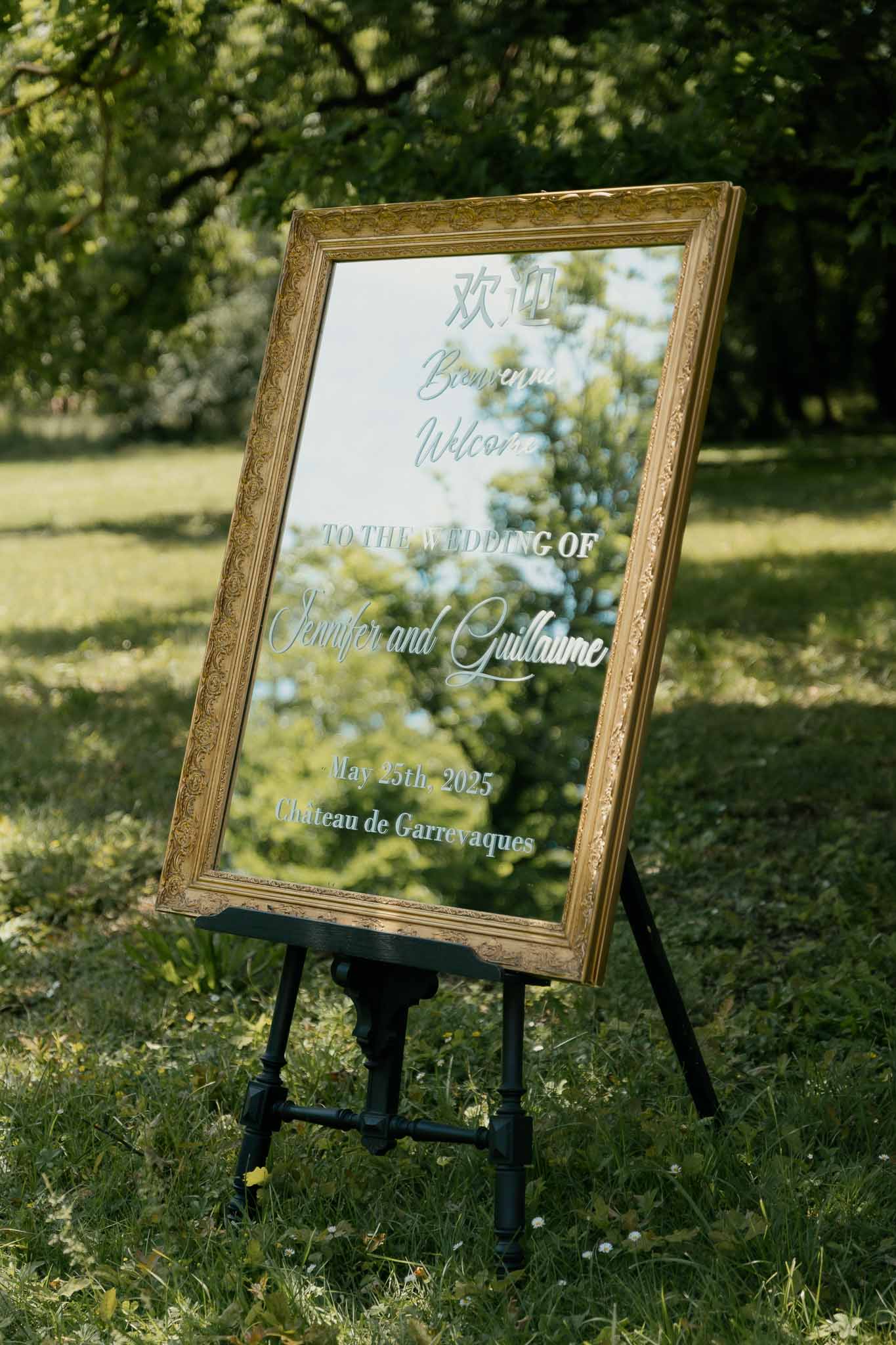 Gold-framed welcome mirror sign with multilingual calligraphy on black easel in garden