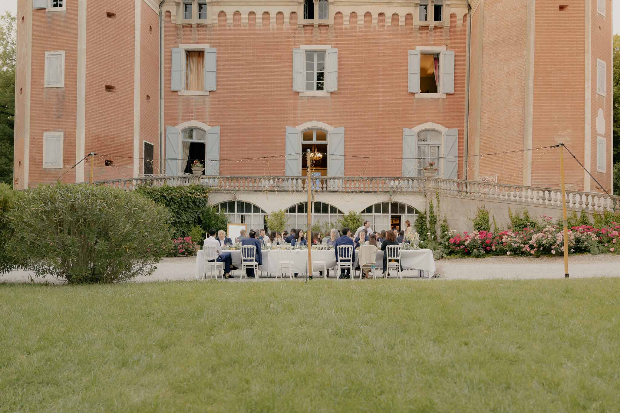 Outdoor reception dinner with guests at long tables before pink brick chateau with fairy lights overhead