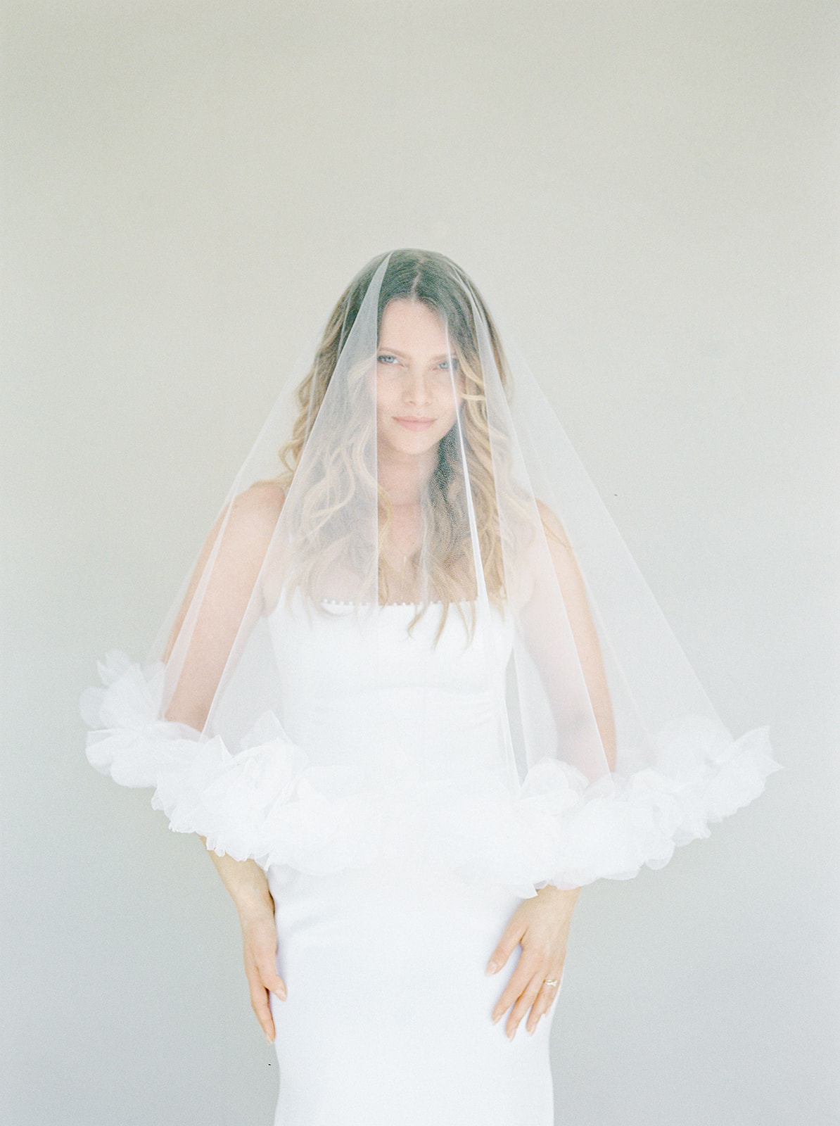 Bride in white strapless gown with organza-edged tulle veil draped over face against grey background